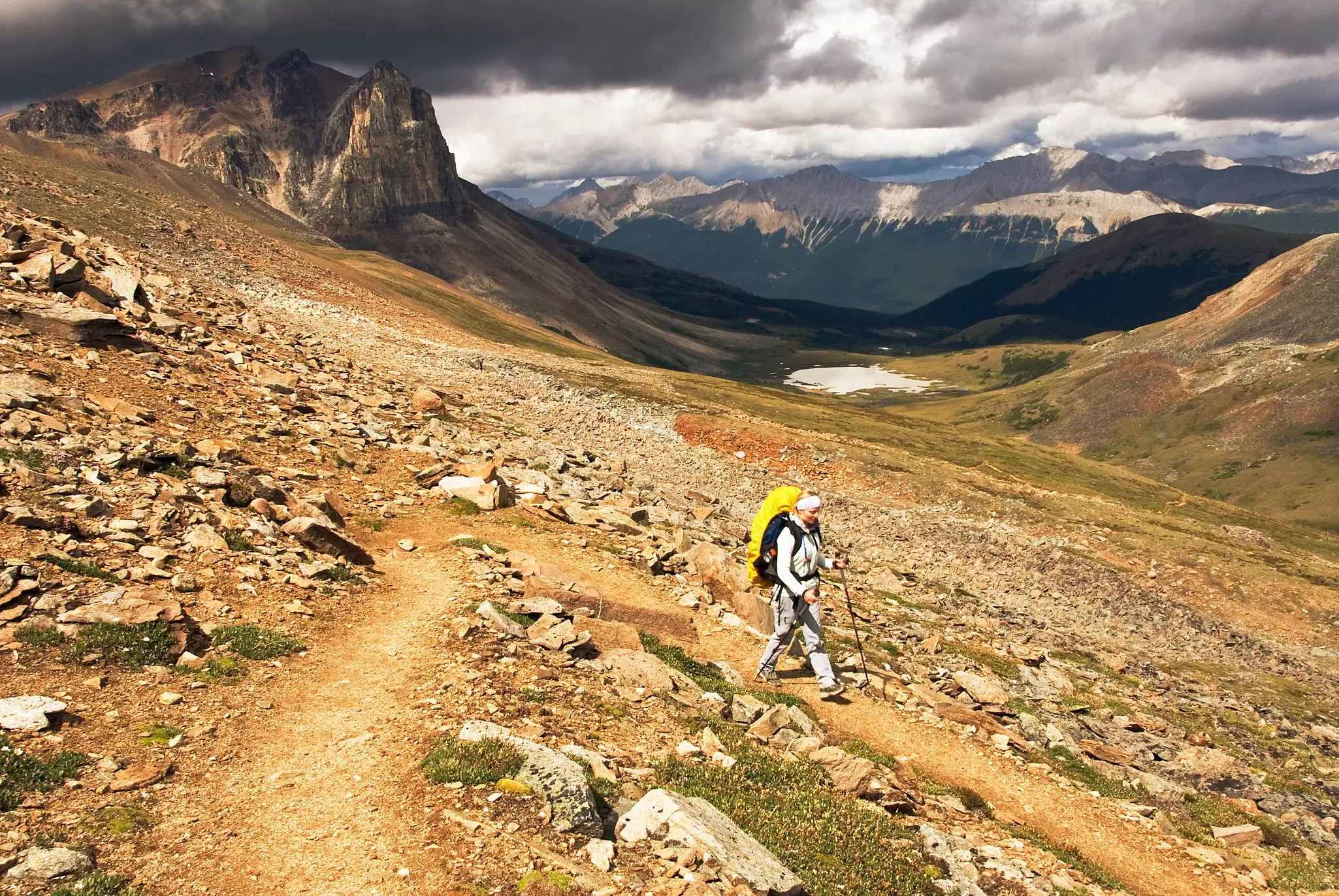 A hiker with a backpack walks a dirt trail across rocky alpine slopes under dark storm clouds and mountain peaks.