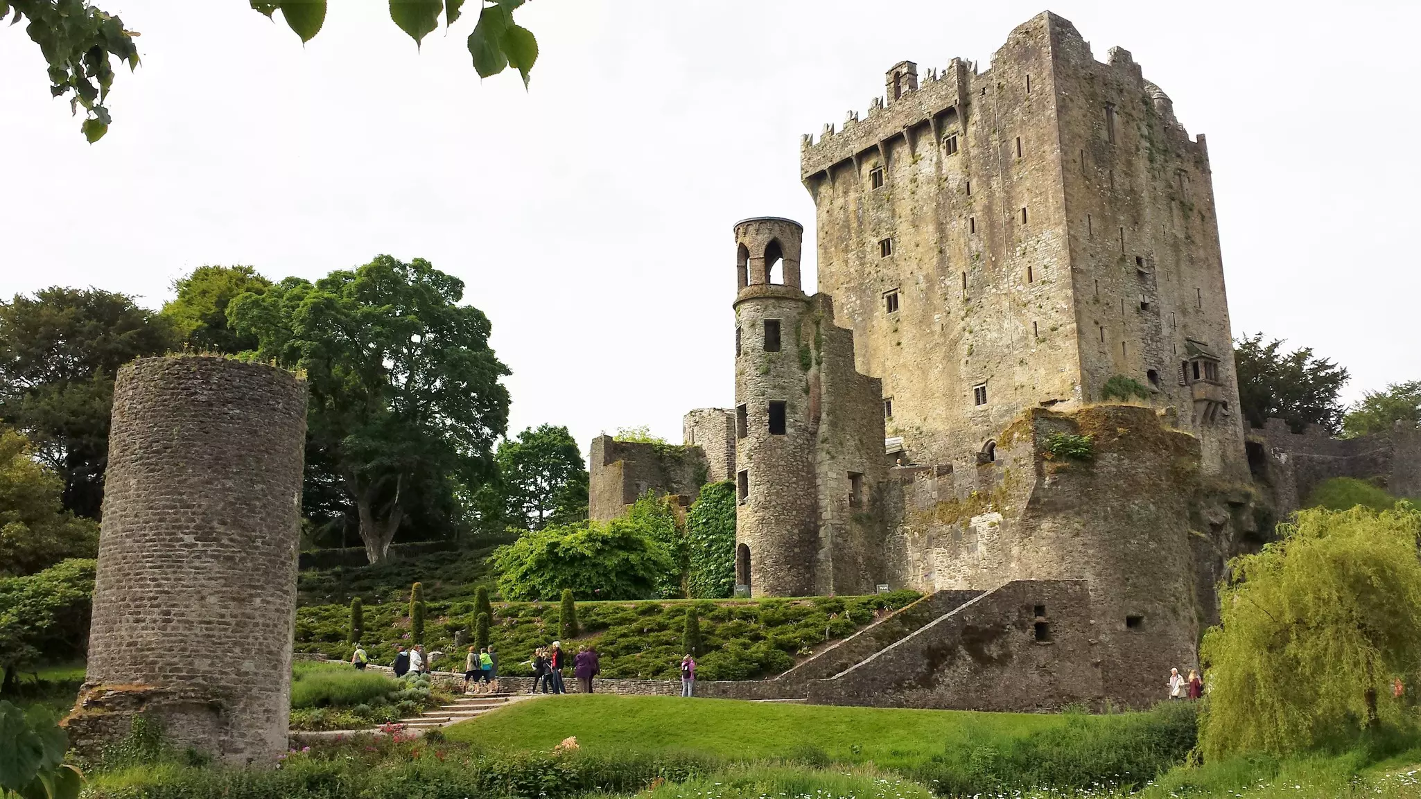 A tarnished castle surrounded by green trees. A few people mill around nearby at the foot of the structure.
