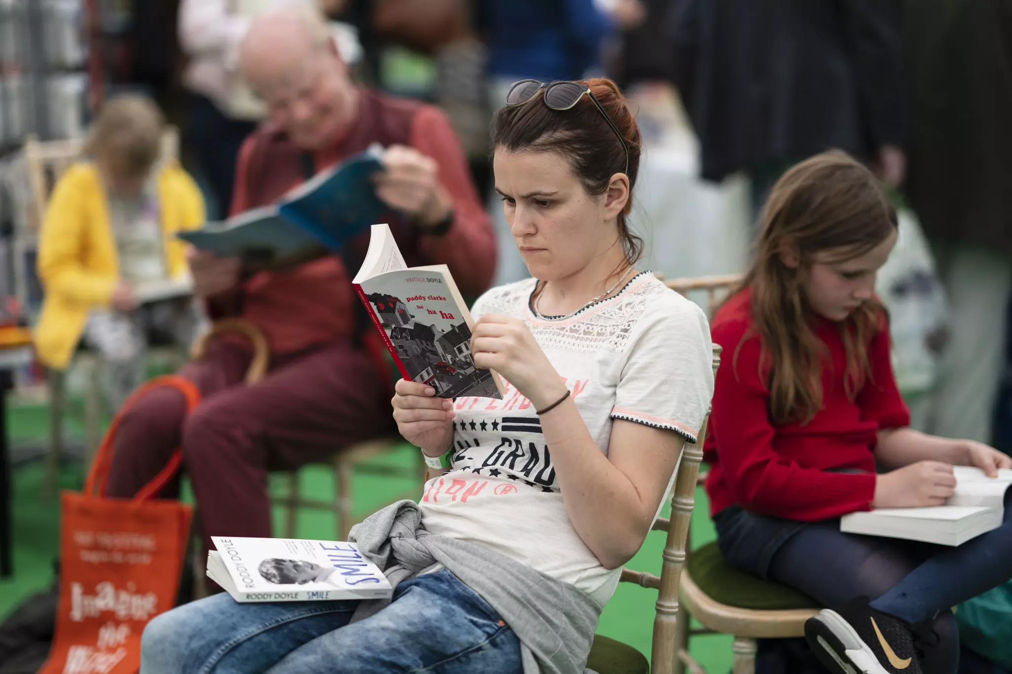 A woman in a t-shirt and other people are seen sitting on chairs and reading books.
