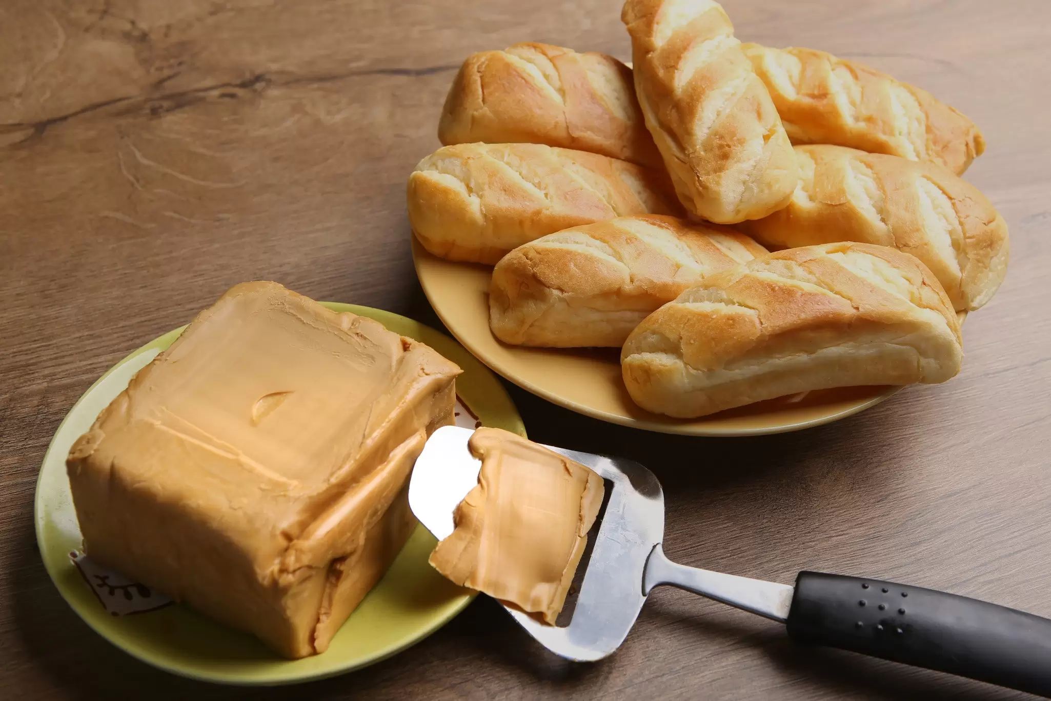 A piece of caramel colored cheese, called brunost, on a wooden table with a slider and some rolls.