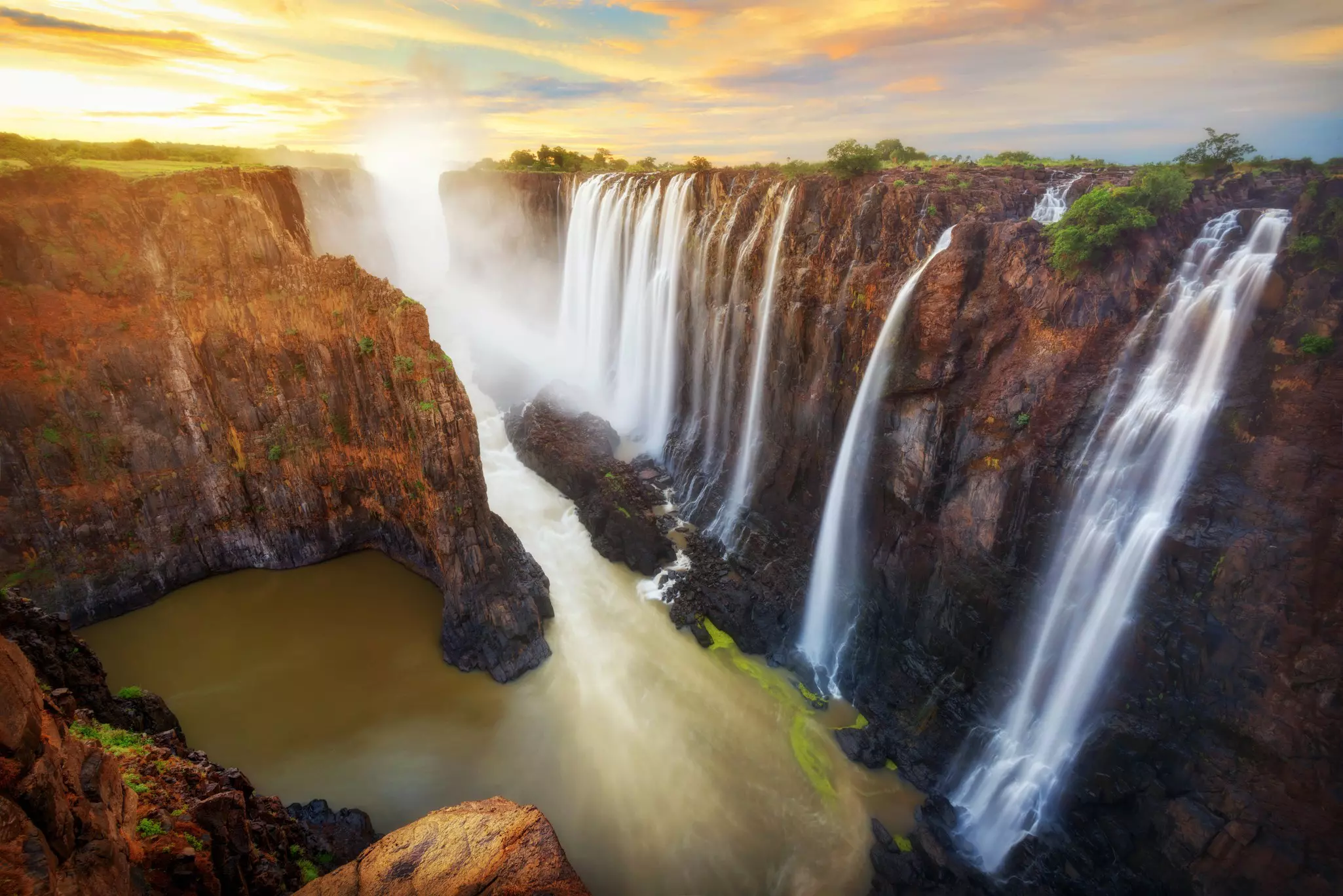 A series of vast waterfalls plunging over rocks that glow red in the sunset.