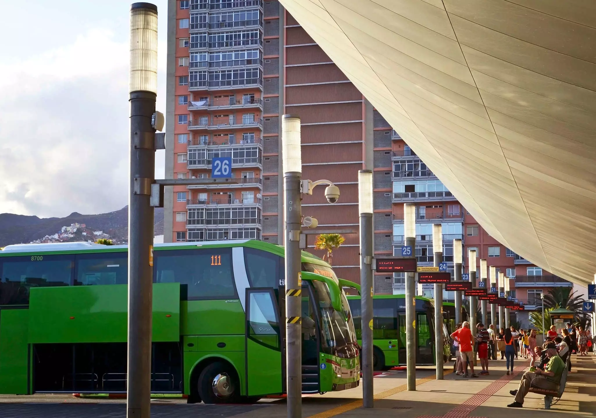 Green buses lined up at an outdoor station on a mostly sunny day. People wait on benches and standing near bus line signs.