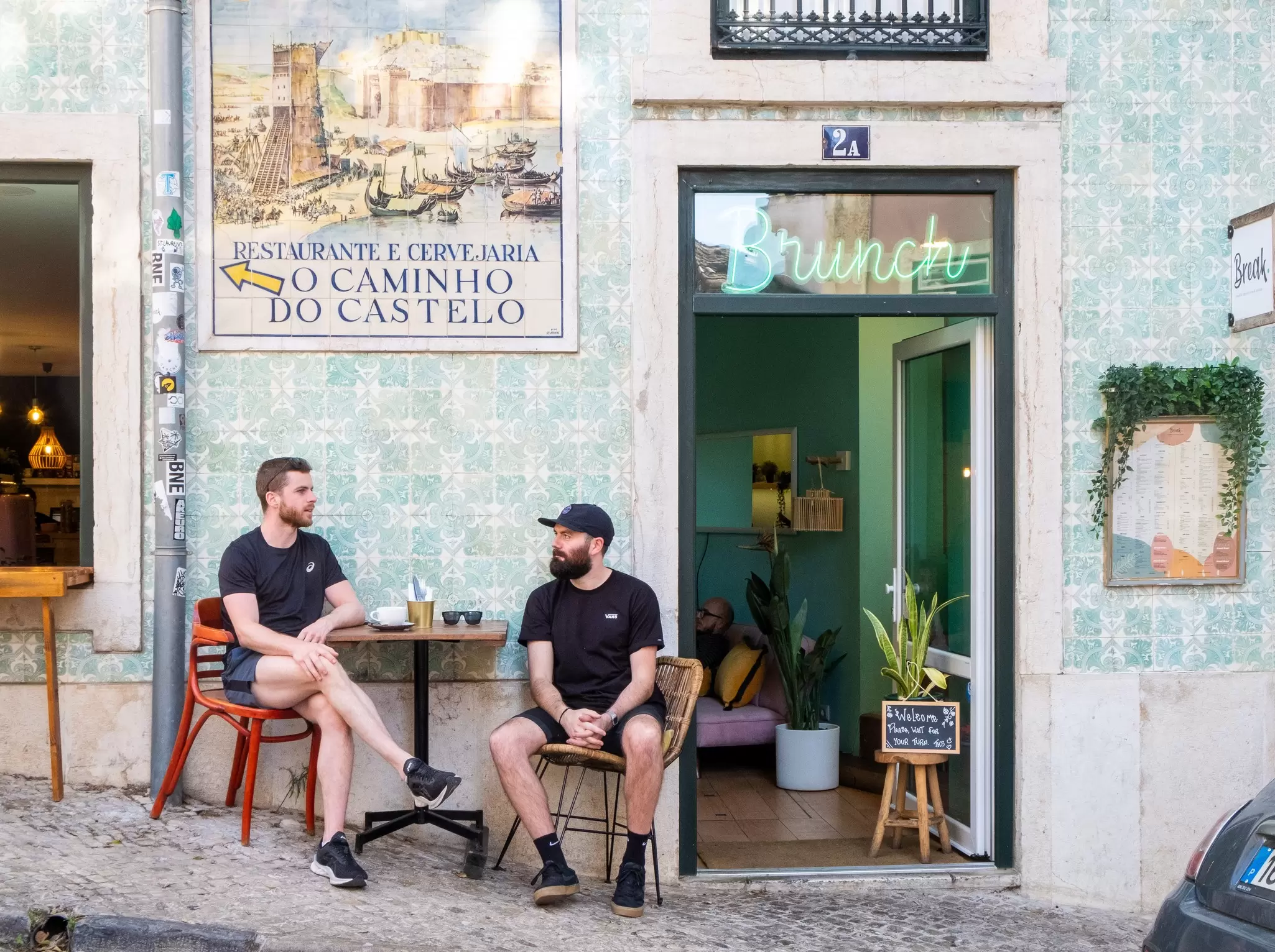 Two people sit at a small table outside a cafe in Lisbon with a blue tiled wall.