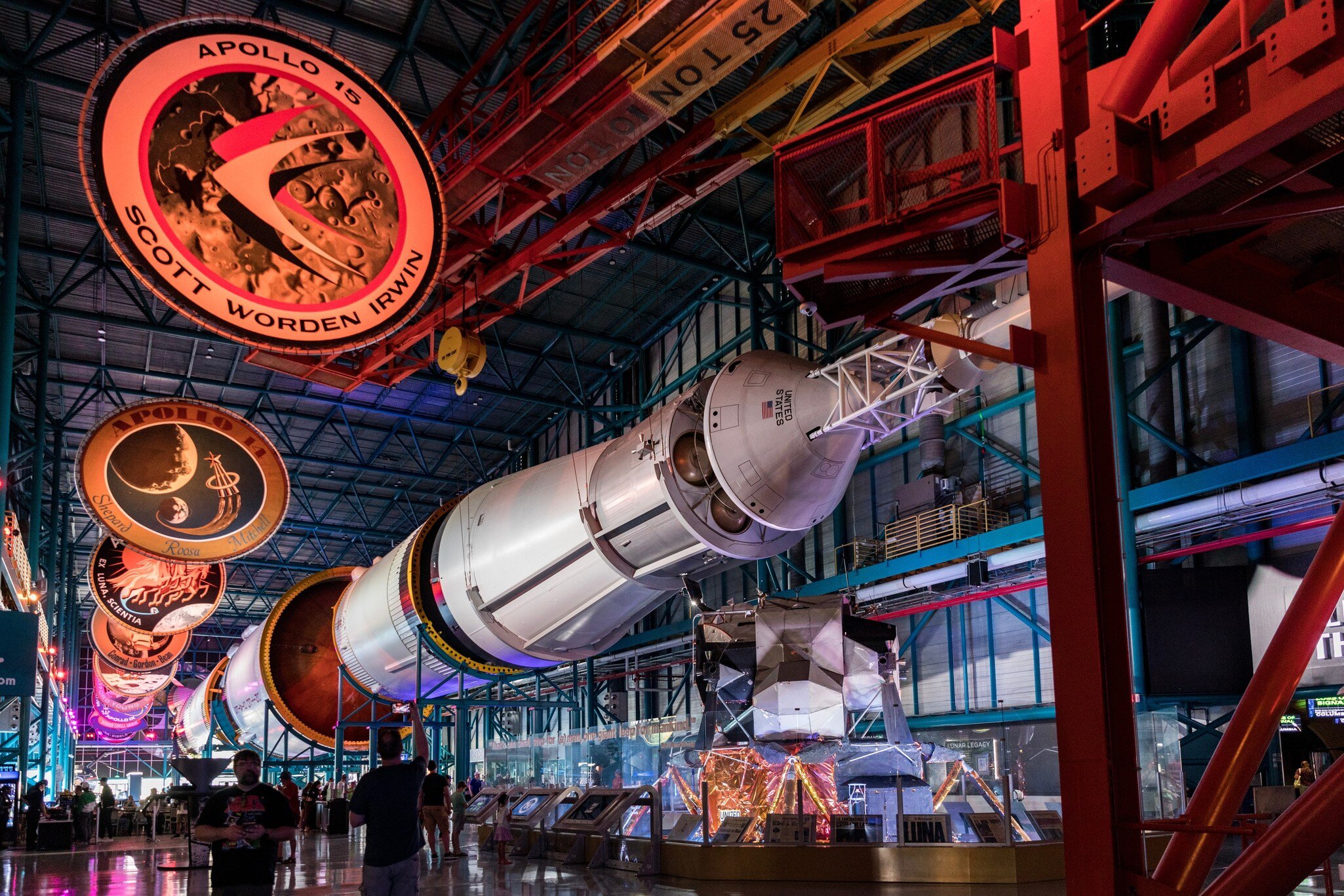 Saturn V rocket lies horizontally under red gantry beams; a hanging disc reads 'Apollo 15 Scott Worden Irwin'.