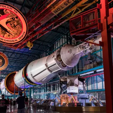 Saturn V rocket lies horizontally under red gantry beams; a hanging disc reads 'Apollo 15 Scott Worden Irwin'.