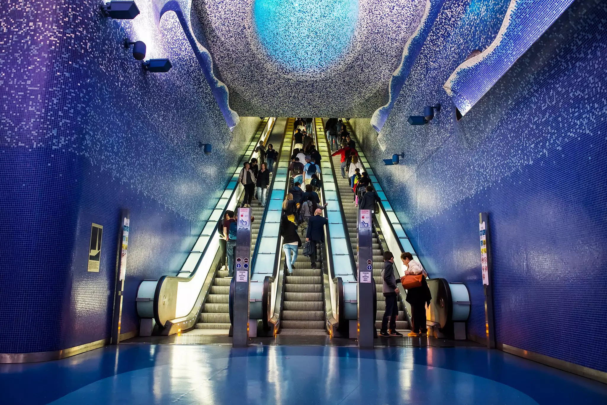 Three escalators rise in a metro station covered with blue and silver mosaic tiles.