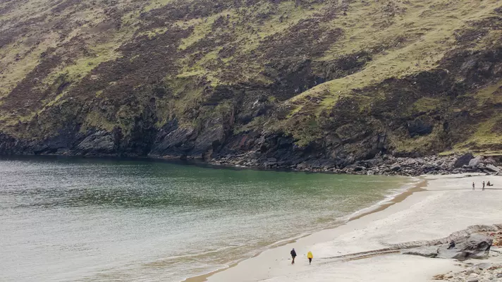 Wide shot of couple in winter gear walking on wide sandy beach with the sea to the left and an imposing, grass-covered tall hill in front of them.