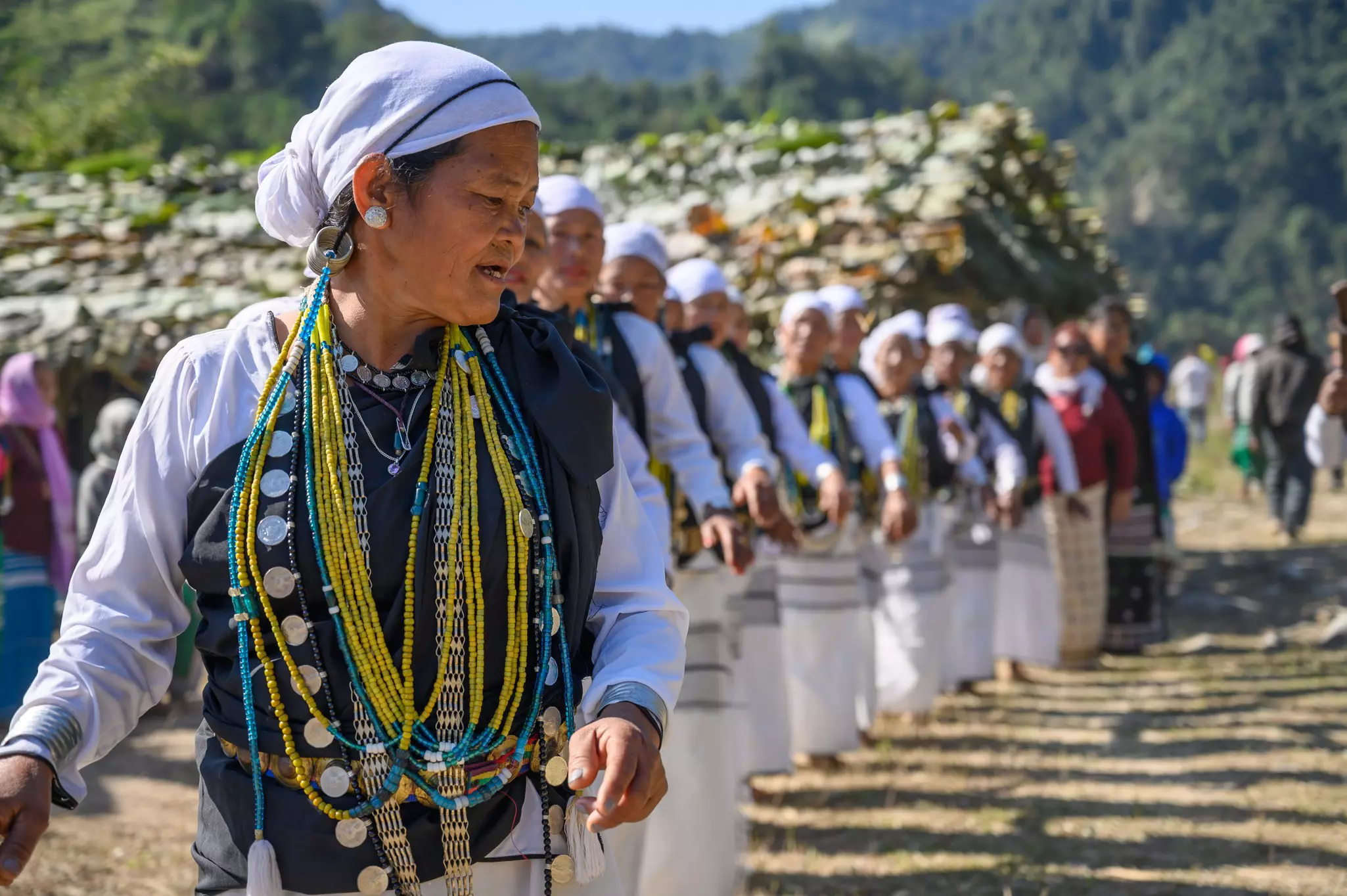 Women in a line making dance movements with their arms. Each is dressed in black and white enhanced by brightly colored beaded necklaces.