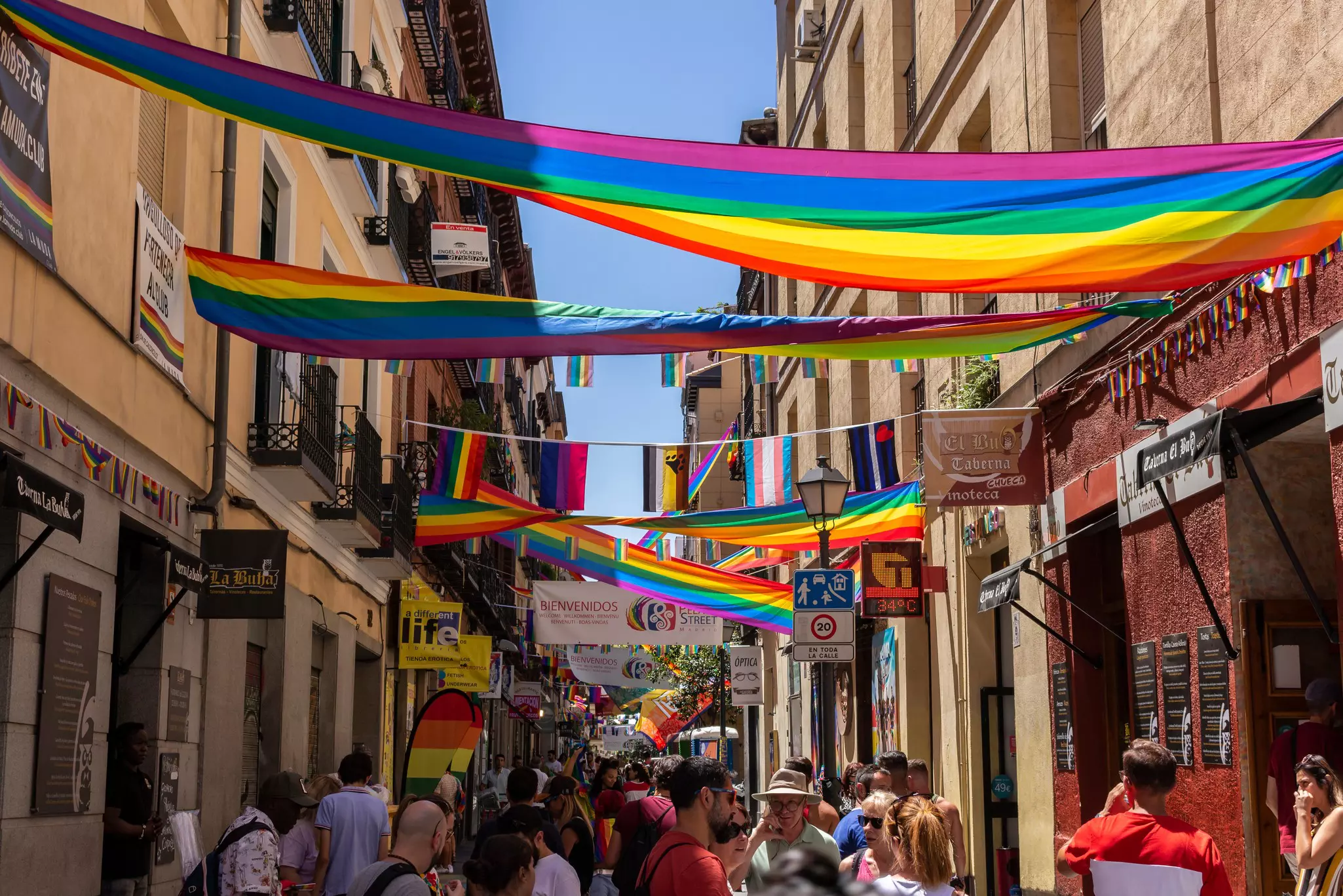 Rainbow flags hang from buildings and over a narrow street in a city neighborhood.
