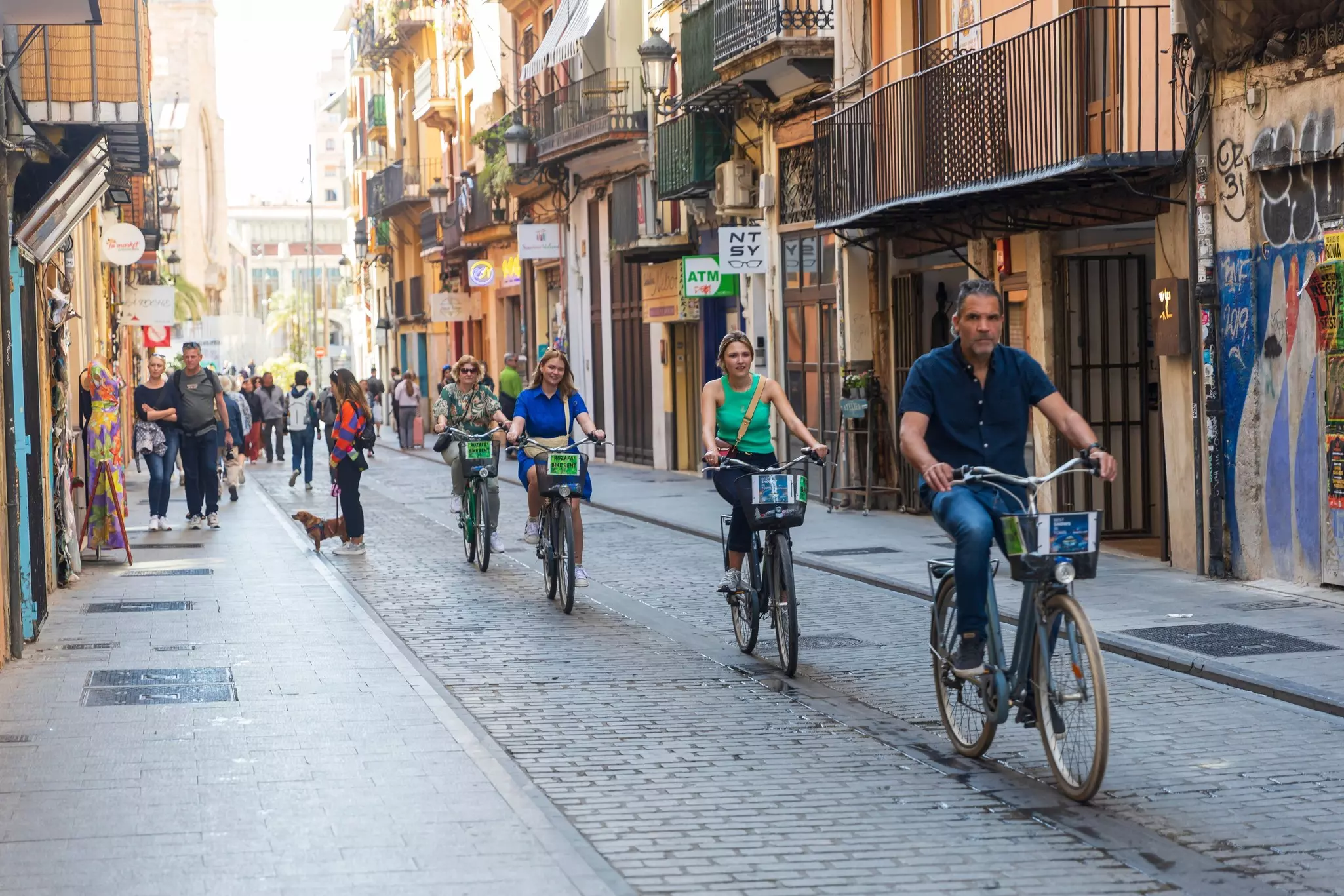 A group of tourists ride rental bikes down a narrow street, exploring the city of Valencia, Spain.