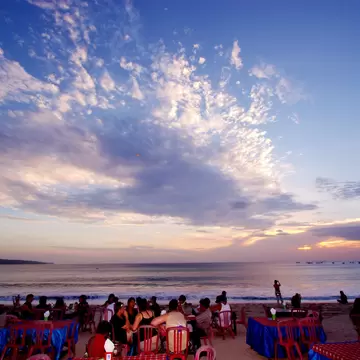 People seated at tables on Jimbaran beach, Bali, under a wide sunset sky streaked with clouds over the sea.