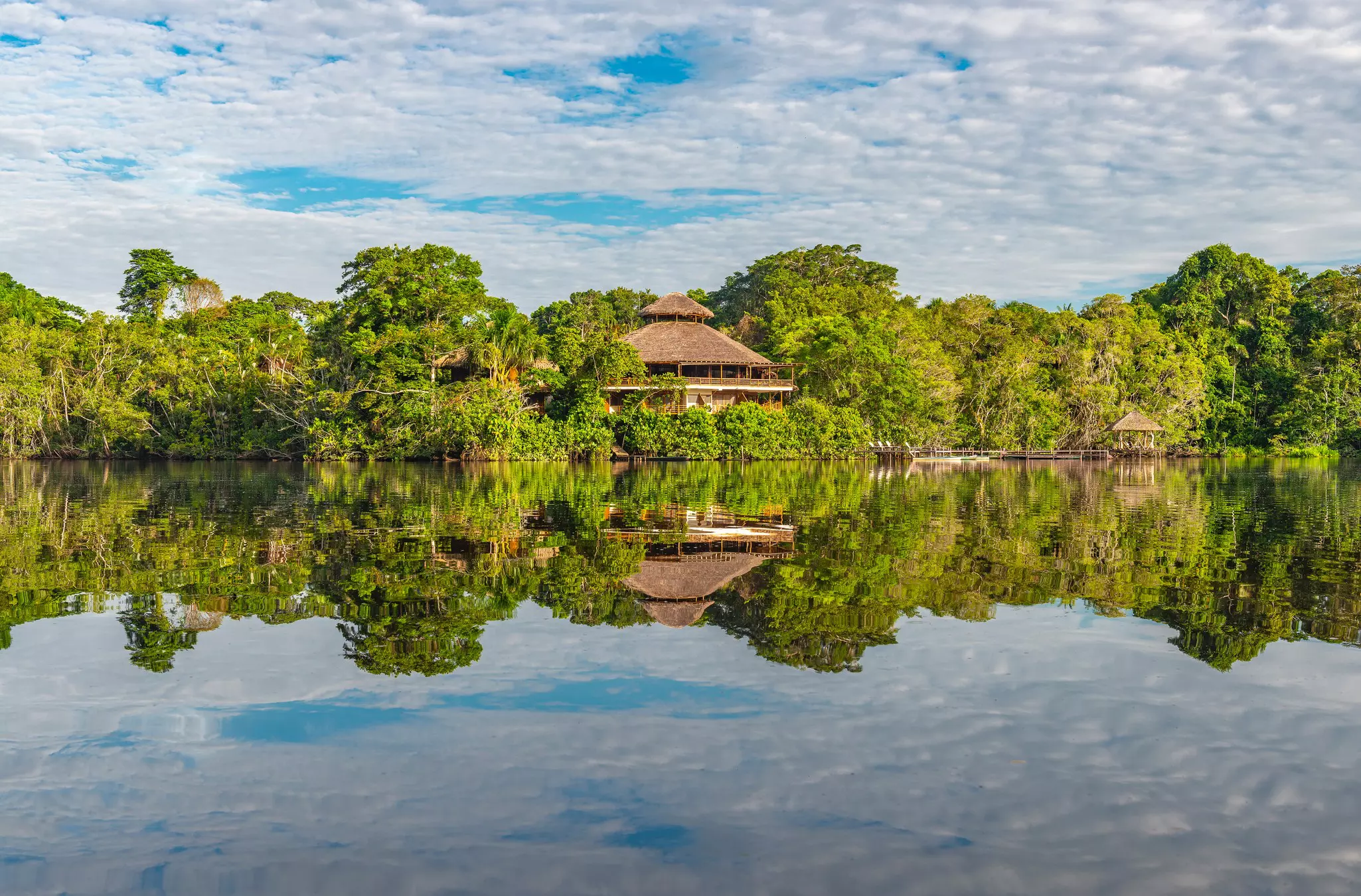 A structure with a conical roof is partially obscured by dense foliage; the building is reflected in water, as are the clouds in the sky above.