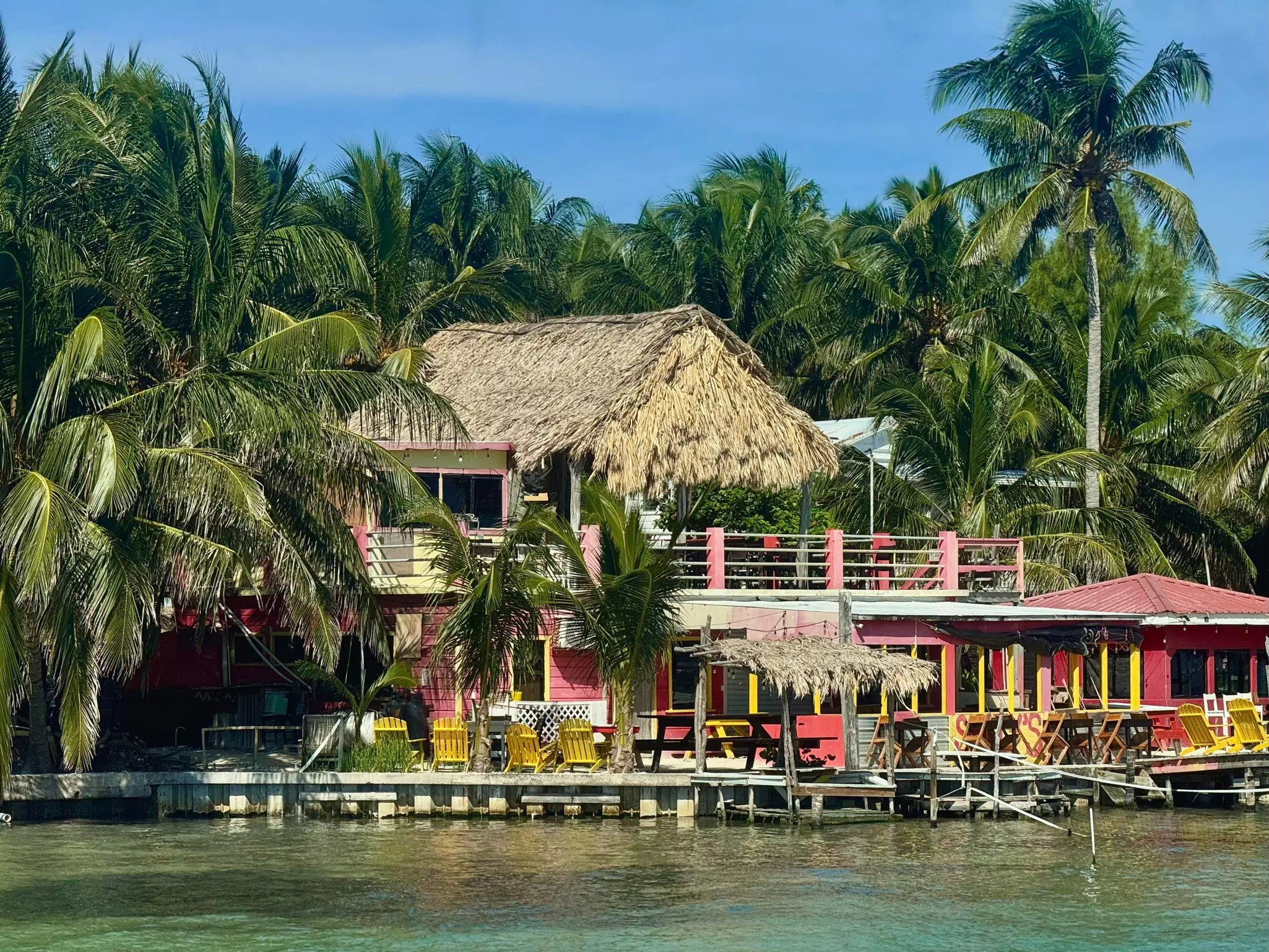 A large pink building by the water. The dock has chairs, and the building has a thatched roof.