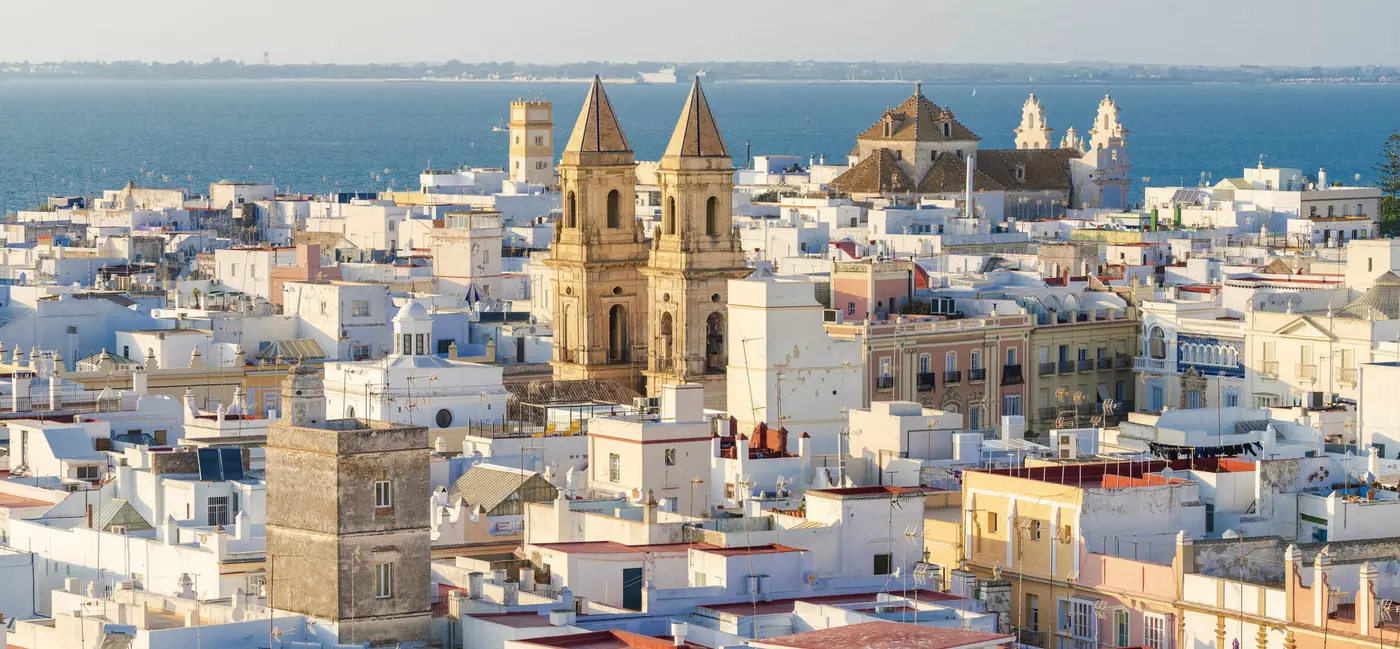A whitewashed, seaside city with low-rise buildings. A cathedral with honey-colored turrets juts out of the middle of the city.