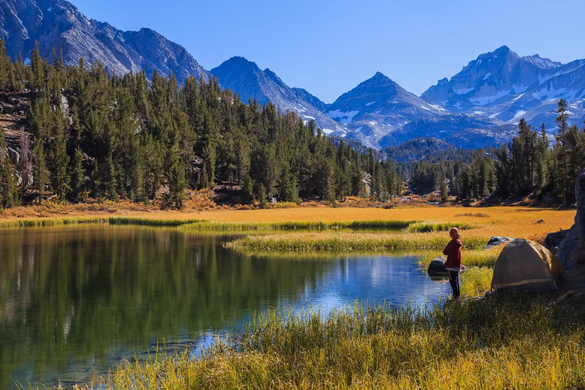 A hiker stands near their tent looking out at lake and mountain scenery that glows gold in the autumn sunlight.