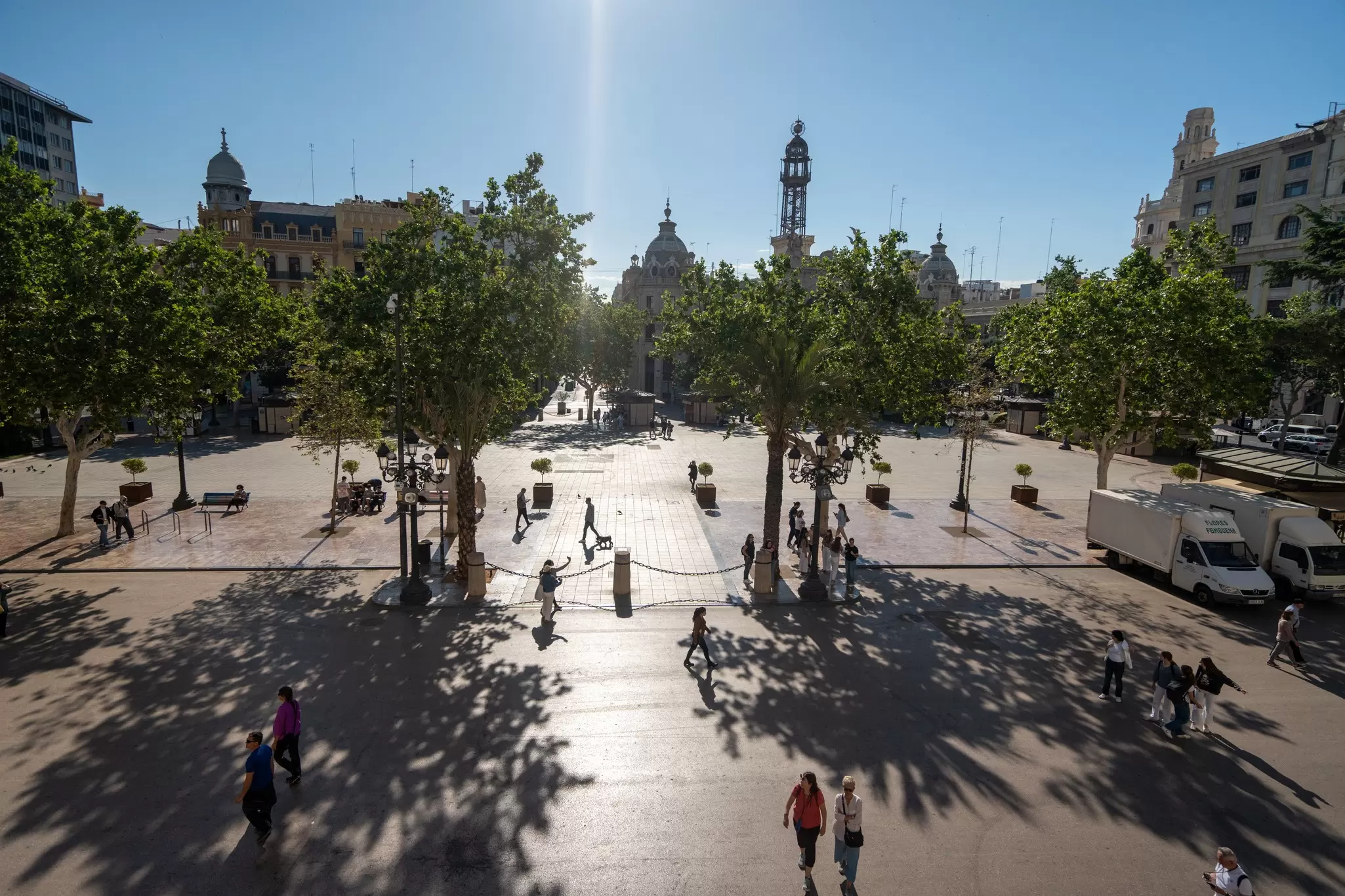 People walk through a large open plaza with some trees and benches.