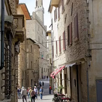 People walking along a narrow stone street in Volterra toward a tall crenellated medieval tower under a pale sky.