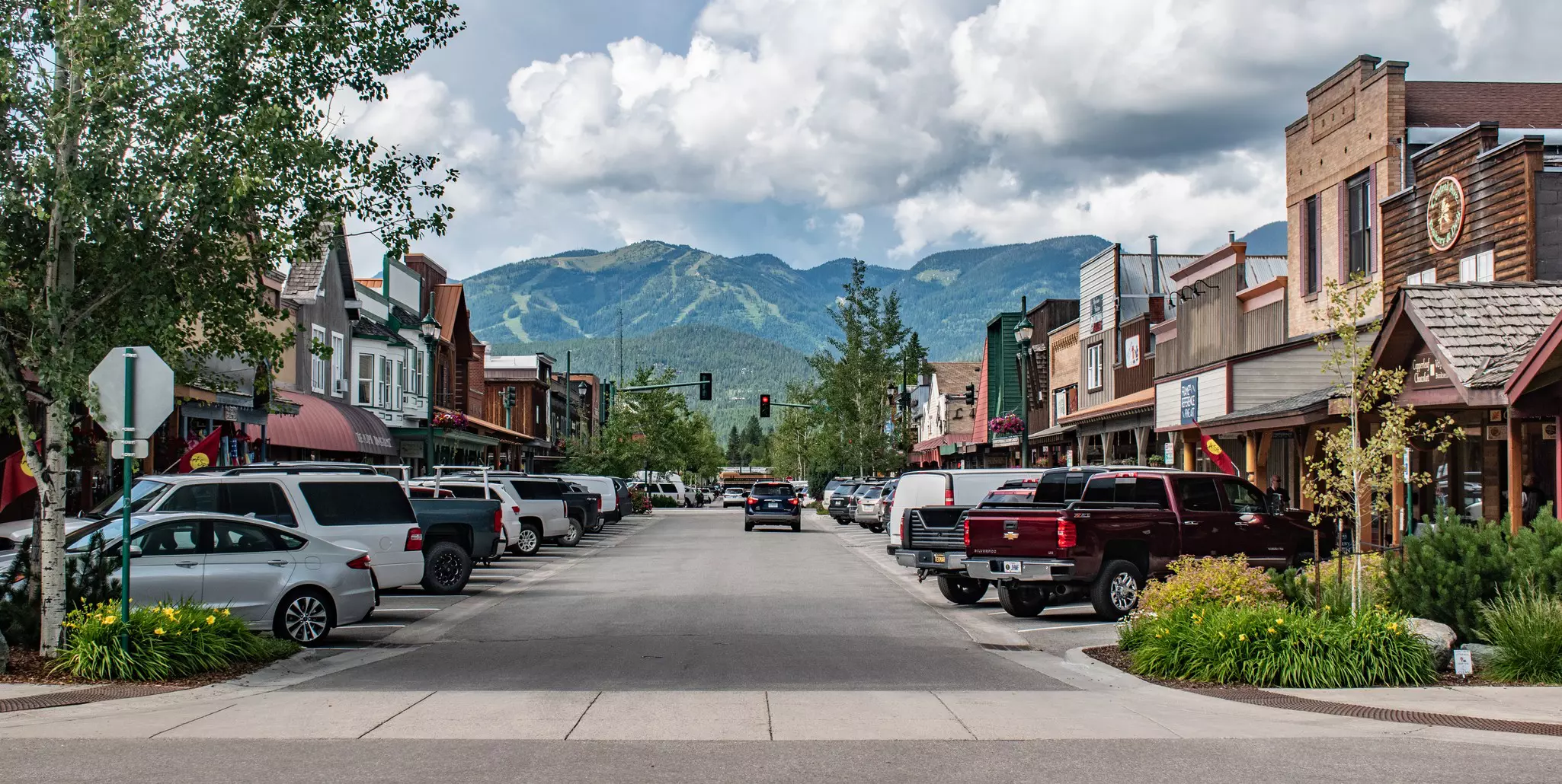 A road with parked cars and low buildings on either side and mountains straight ahead in the background.