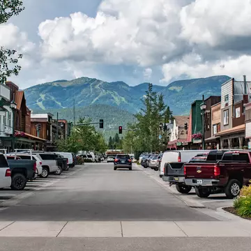 A road with parked cars and low buildings on either side and mountains straight ahead in the background.