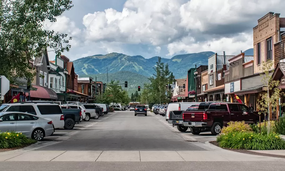 A road with parked cars and low buildings on either side and mountains straight ahead in the background.