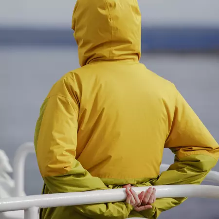 A person in a yellow waterproof jacket stands on a ferry, grasping a white railing.