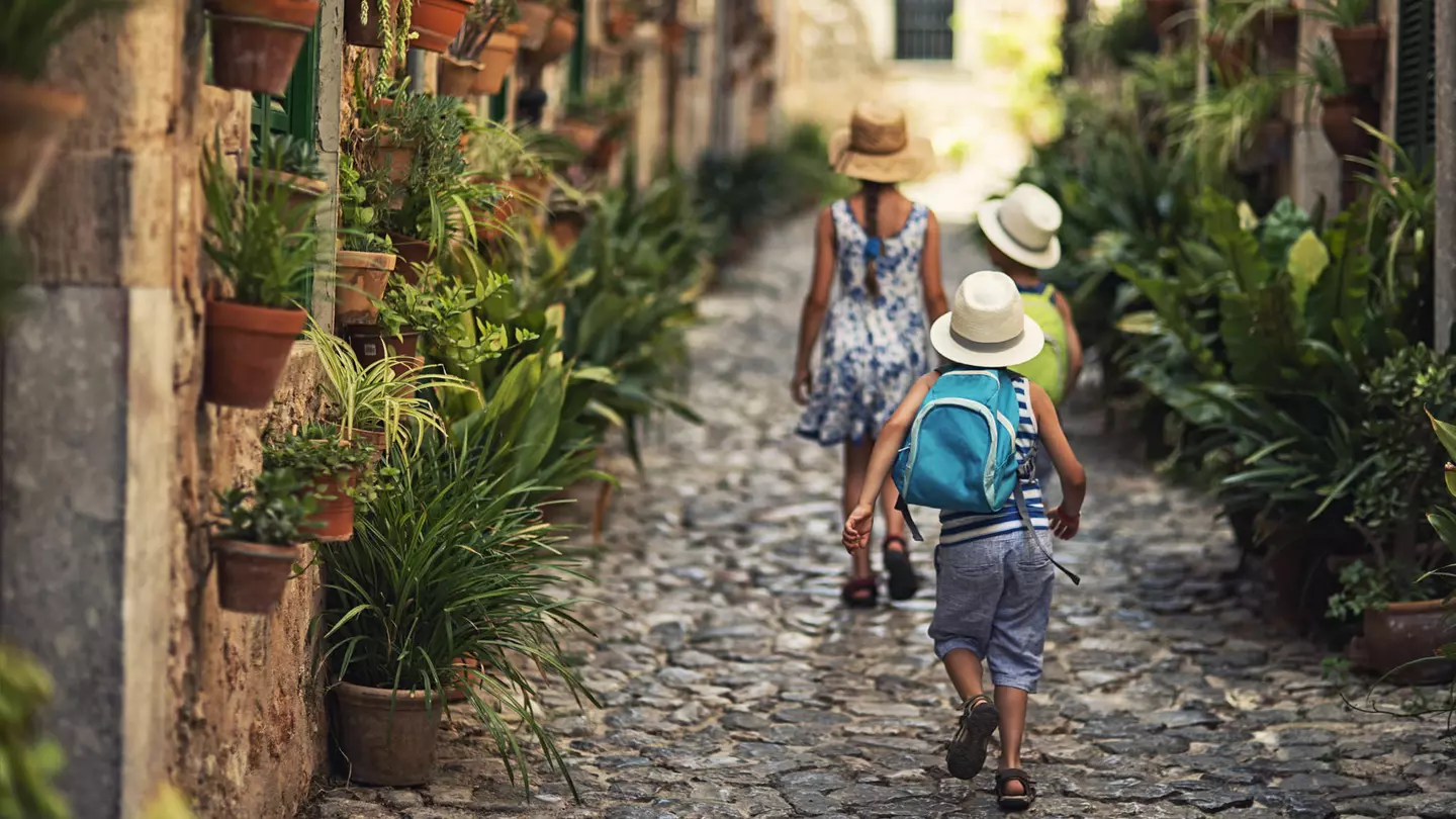 Three kids walking away from the camera in a cobble-stone alley lined with potted plants; the kids are all wearing sunhats and two have backpacks
