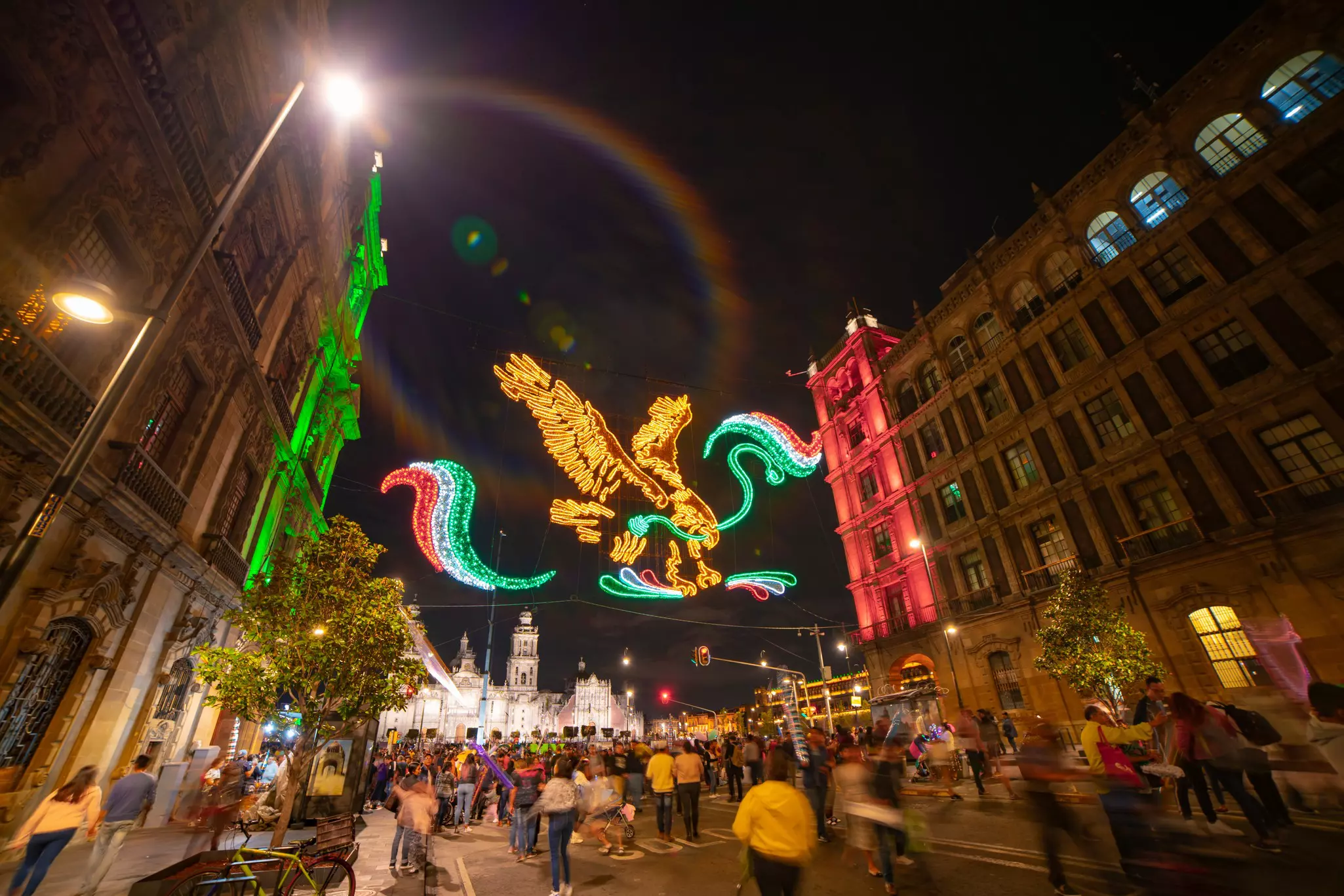 An eagle in colorful neon lights strung across a city street at night, surrounded by buildings with red and green lights and out-of-focus people walking on the street.