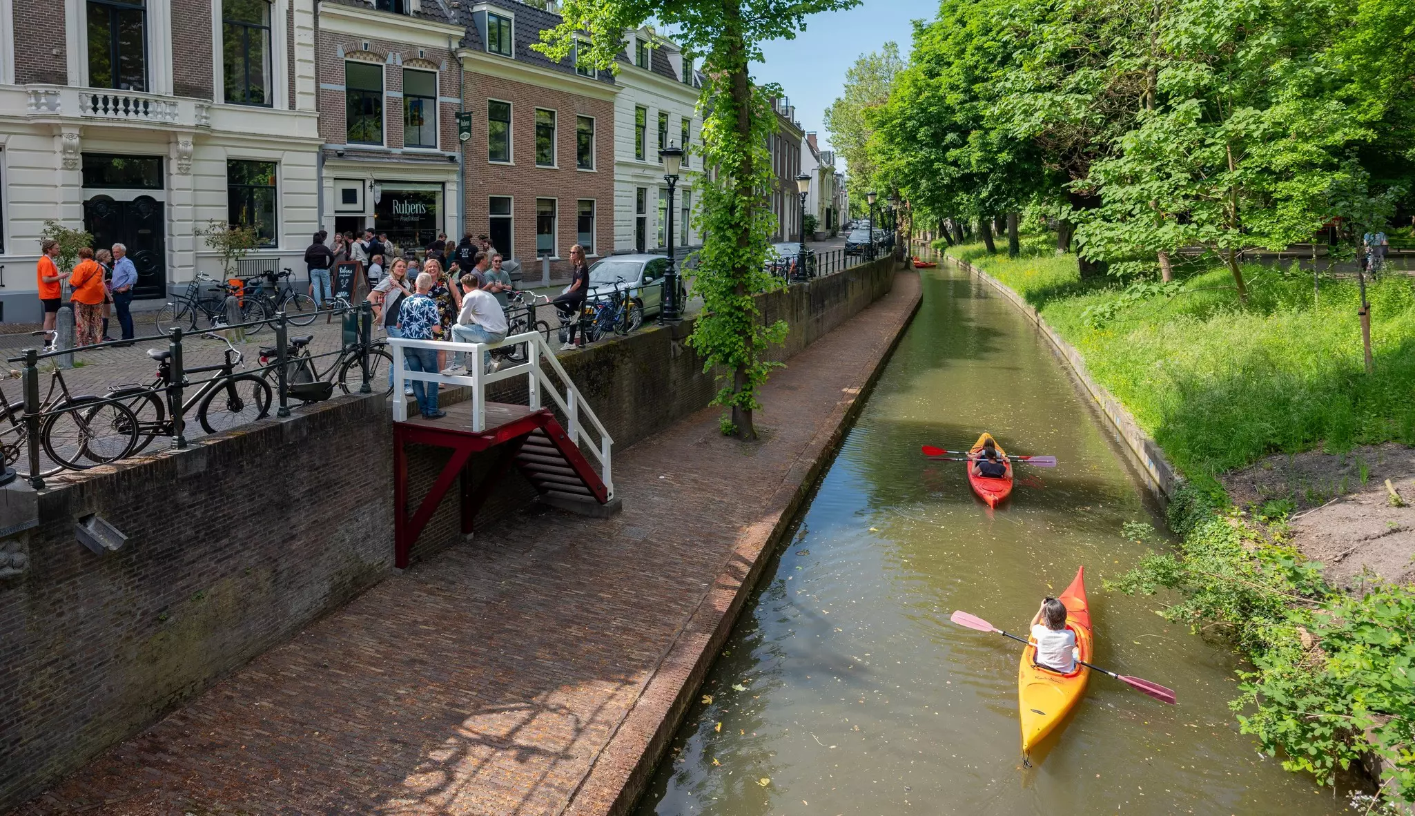 Two kayakers paddle on a narrow canal in a city; people, parked cars and bicycles are on the street that runs parallel to the canal.