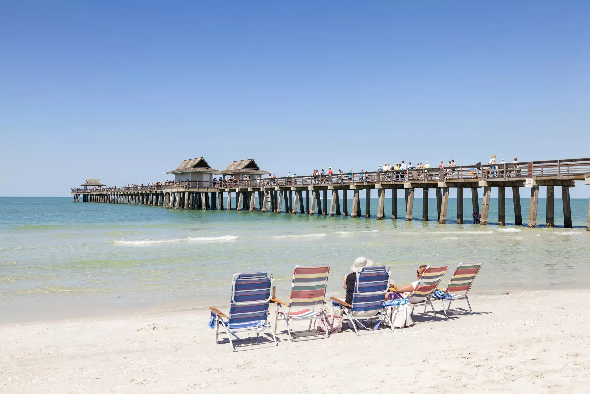 A row of deckchairs on a white sand beach. People walk along the nearby wooden pier.