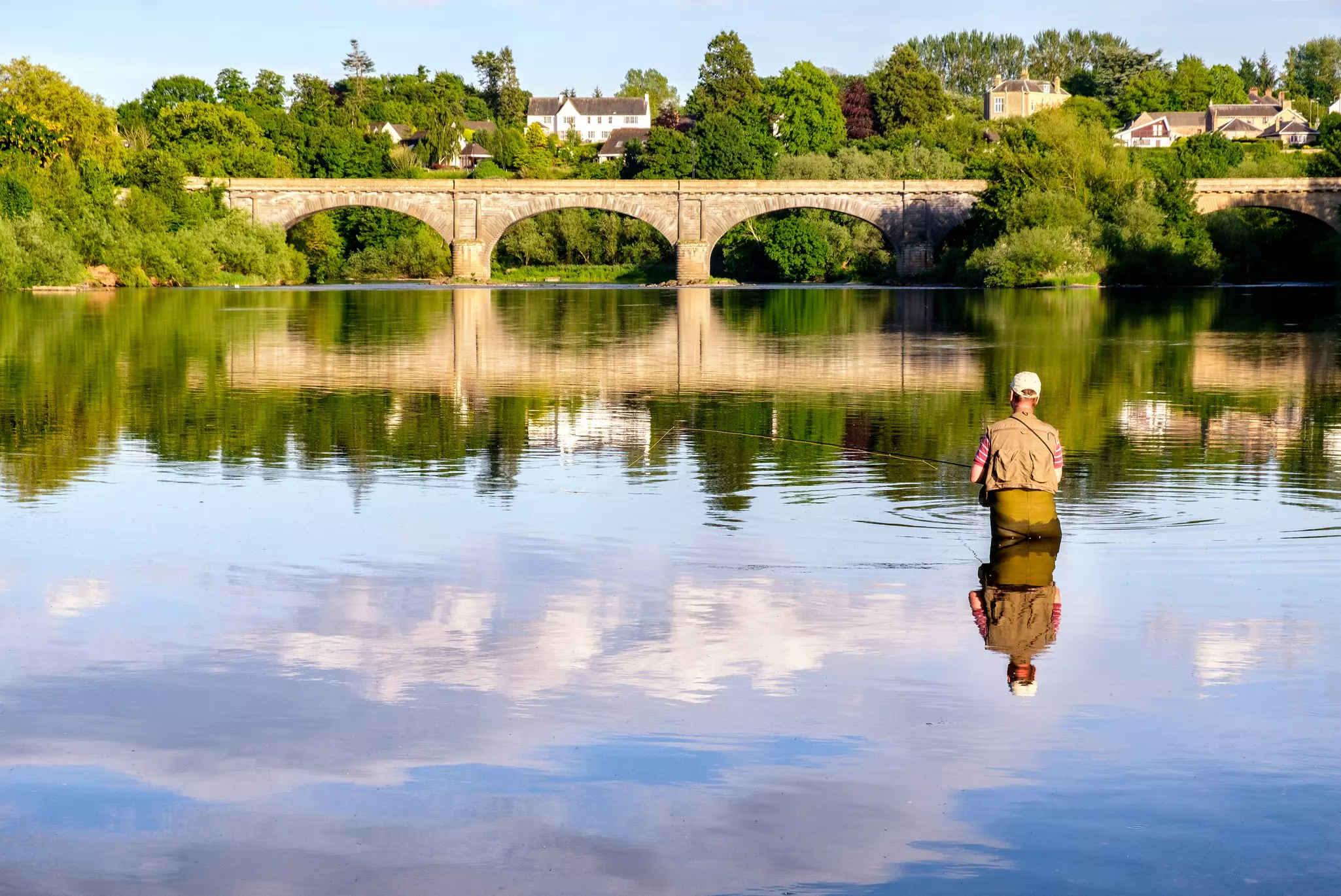 A man stands in a river up to his waist as he casts a fishing line. A stone bridge with many arches crosses the river nearby.