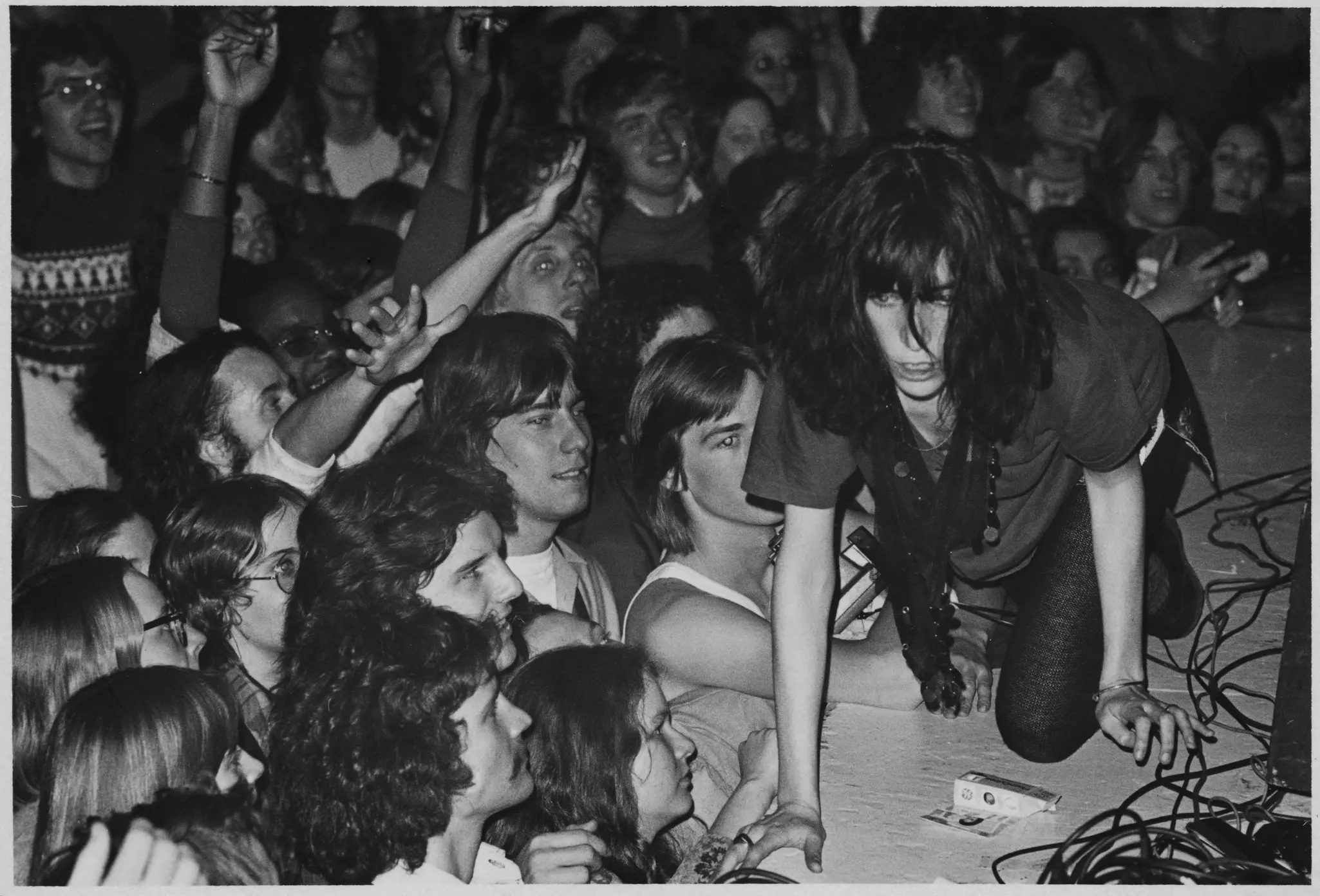 A black and white photo of a woman crawling a cross a stage as the audience cheers and reaches for her.