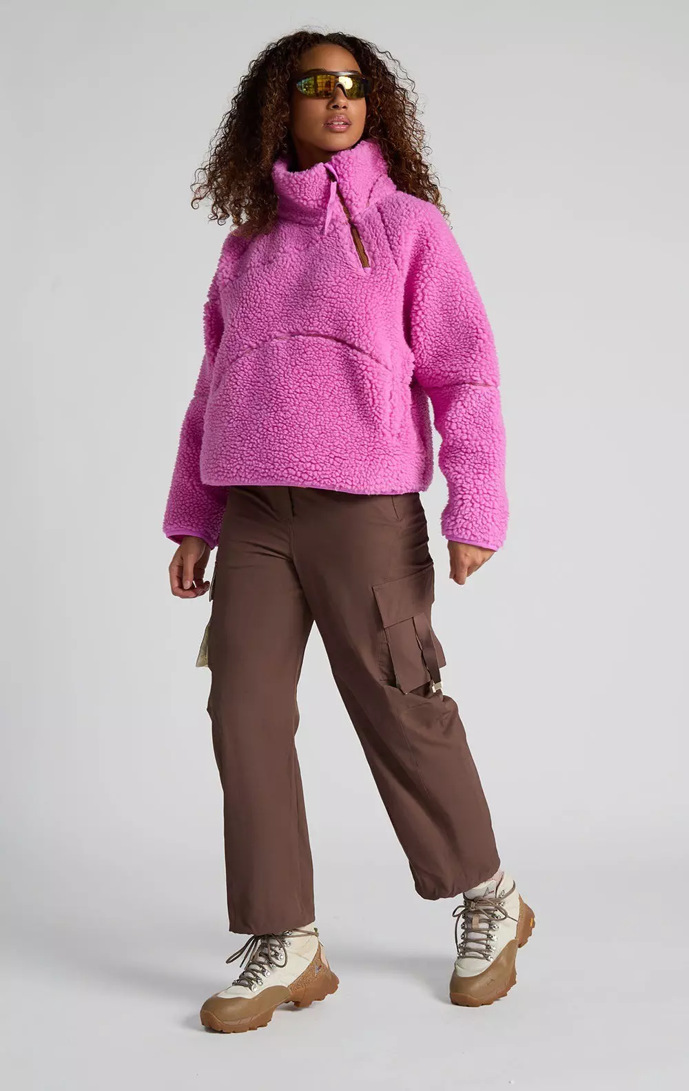 A model in a pink fleece top, brown pants, and white and tan hiking boots.