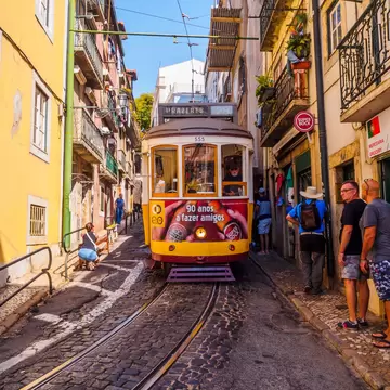 A yellow-painted tram with ads is seem from the front rolling down a narrow, cobbled street on a hill in a city.