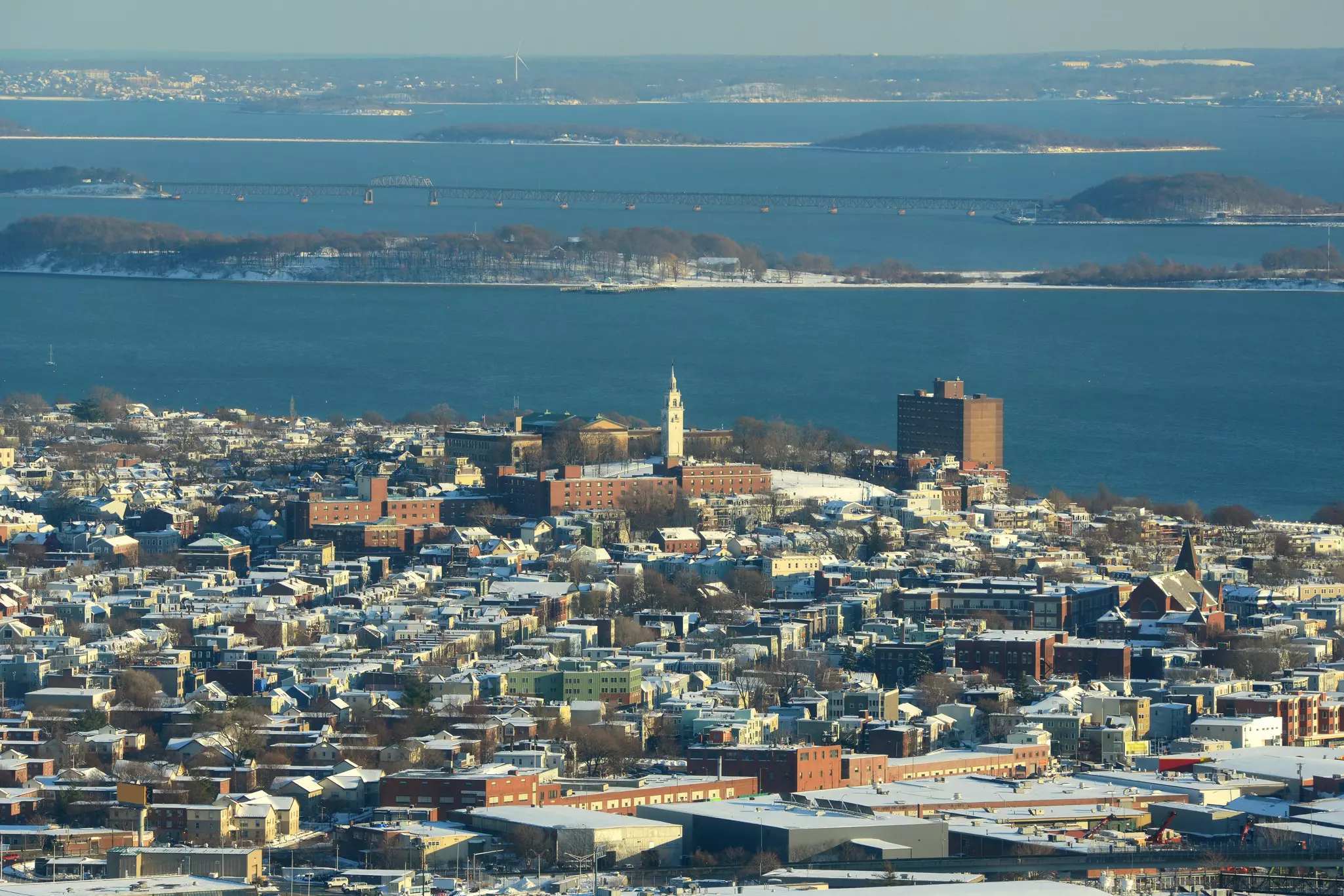 A seaside city neighborhood on a winter's day, with snow covereding the roofs and opens spaces.