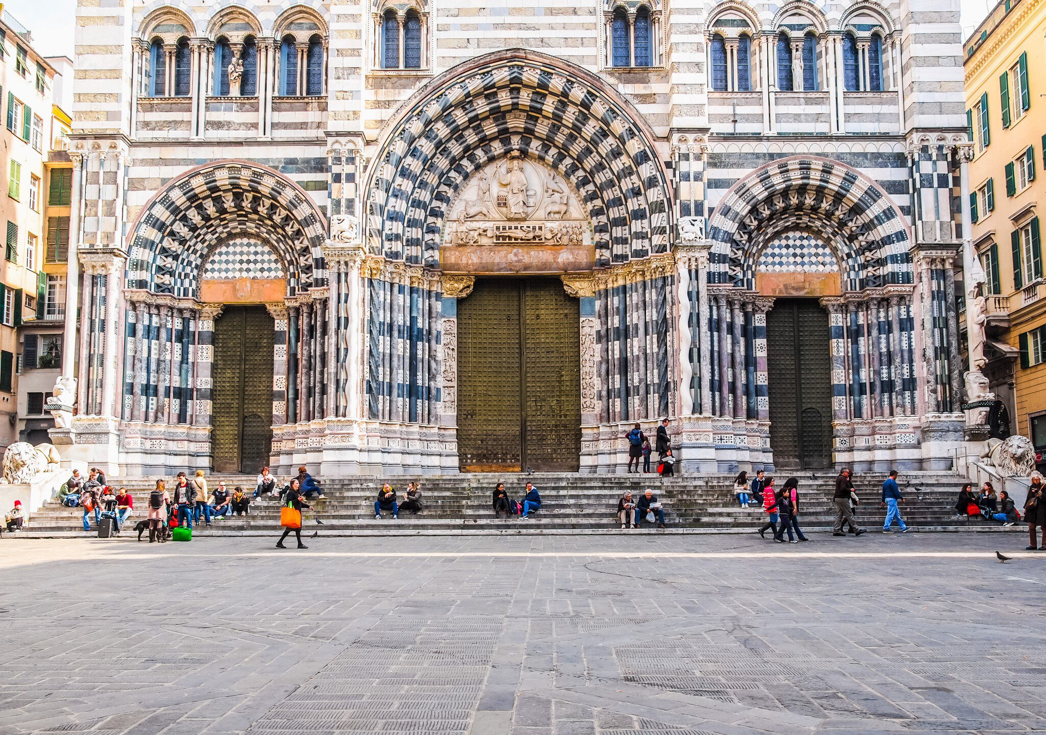 People sit on the steps at the front of a Gothic and Romanesque-style cathedral.