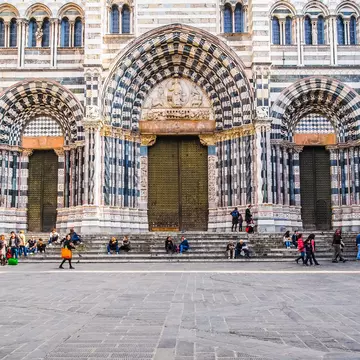 People sit on the steps at the front of a Gothic and Romanesque-style cathedral.