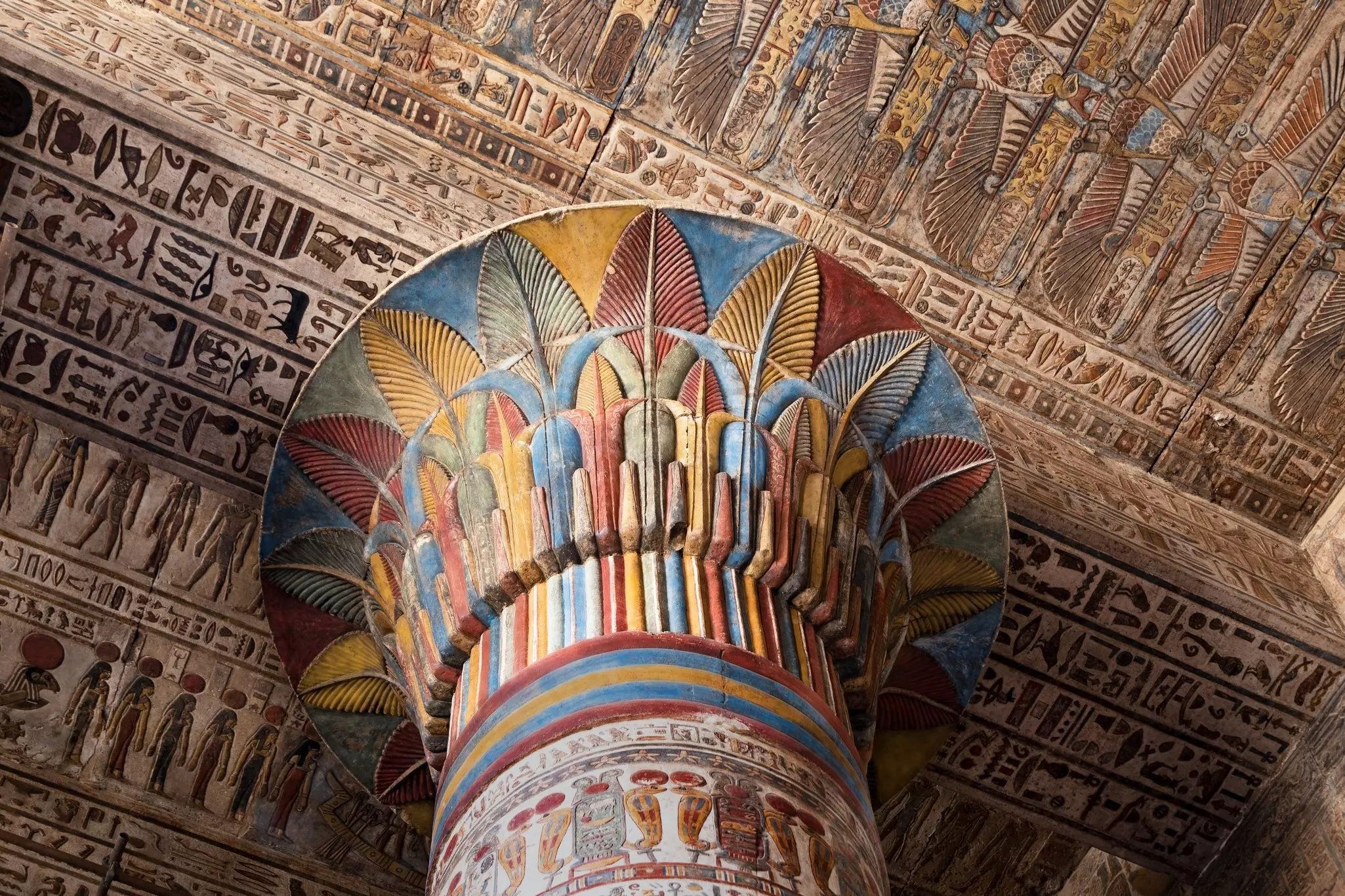 A tall column covered in engravings and markings in yellow, blue and red meets a heavily decorated ceiling.