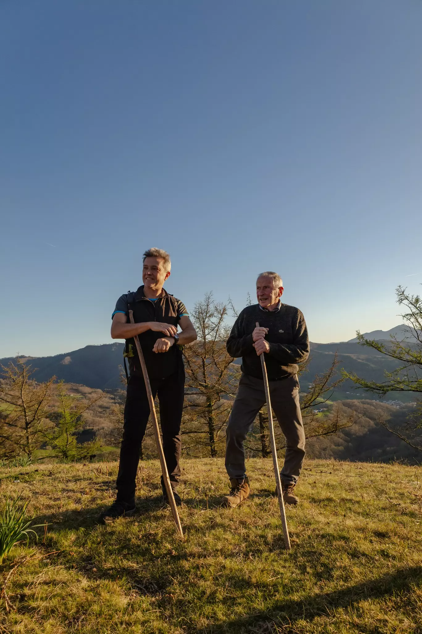 Two men stand with walking sticks in a mountainous area.