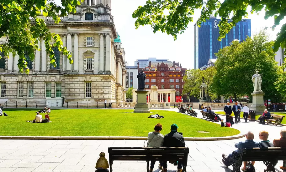 People relaxing on the sunny lawns of a grand columned civic building, with statues, benches and city towers behind
