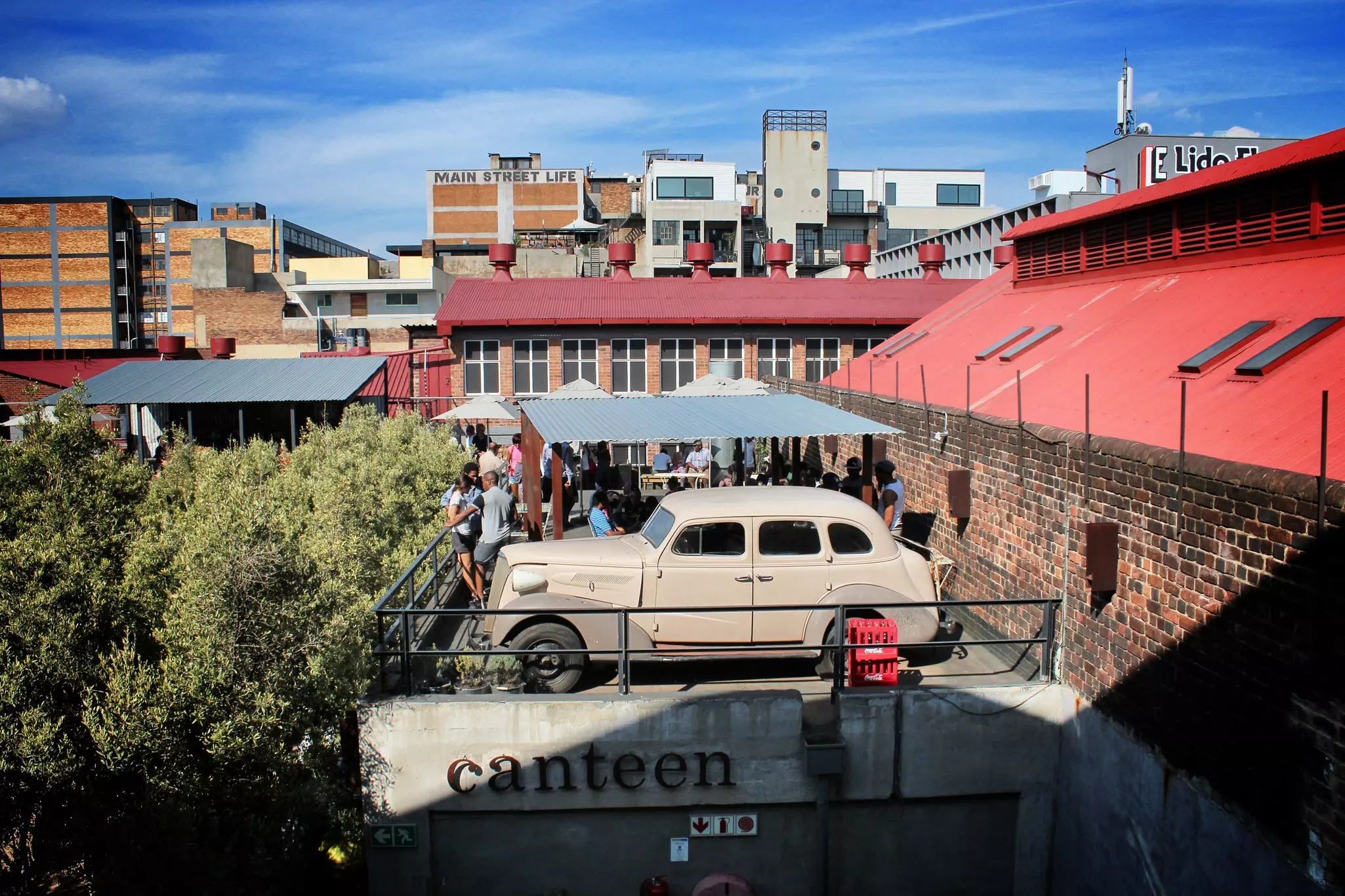 A crowd gathers under an awning on the flat roof of Market on Main, sharing the space is a vintage 1930s car. Trees climb up to this level on the left and renovated old industrial buildings back the scene.