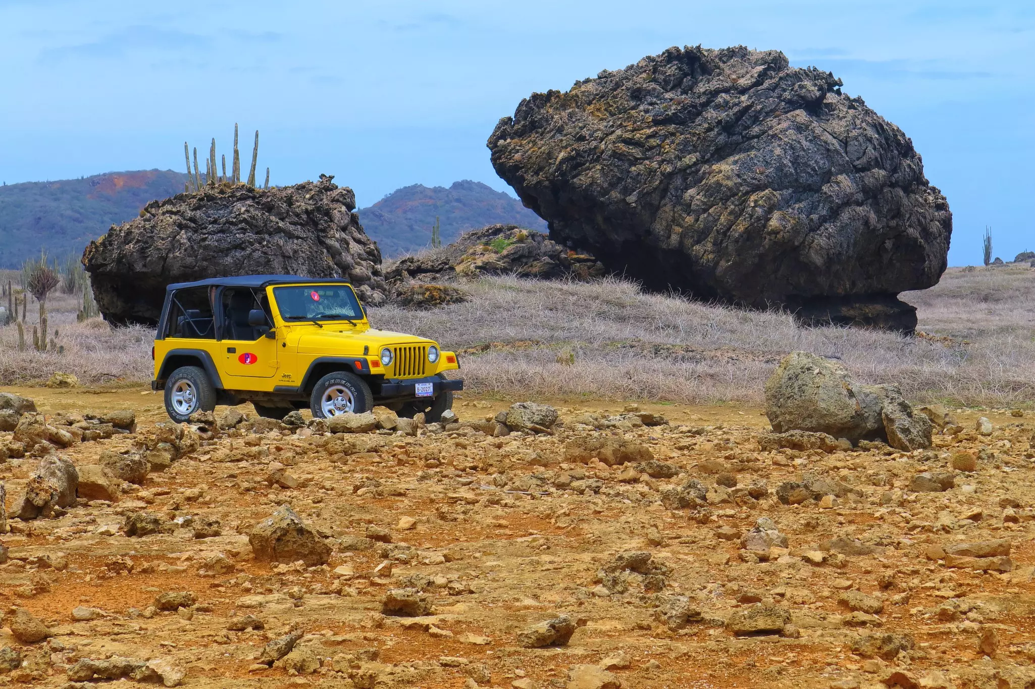 A yellow jeep drives through a dusty landscape past huge boulders.