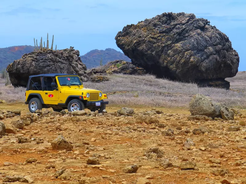A yellow jeep drives through a dusty landscape past huge boulders.
