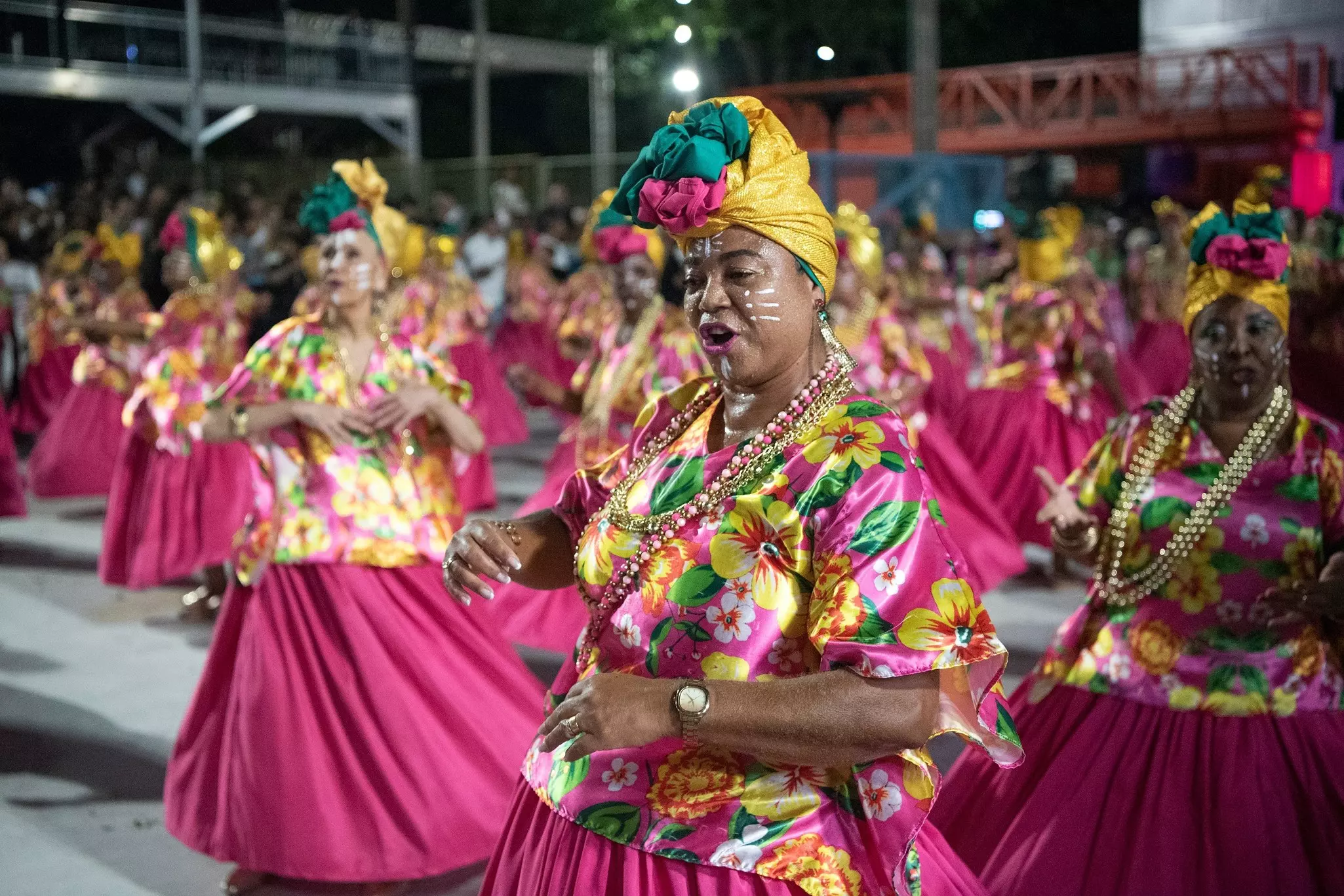 Women wearing bright pink, yellow and green floral outfits at a technical rehearsal for Carnaval