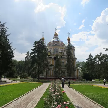 Yellow cathedral with patterned domes behind pine trees, twin paved paths and a rose bed leading towards it.