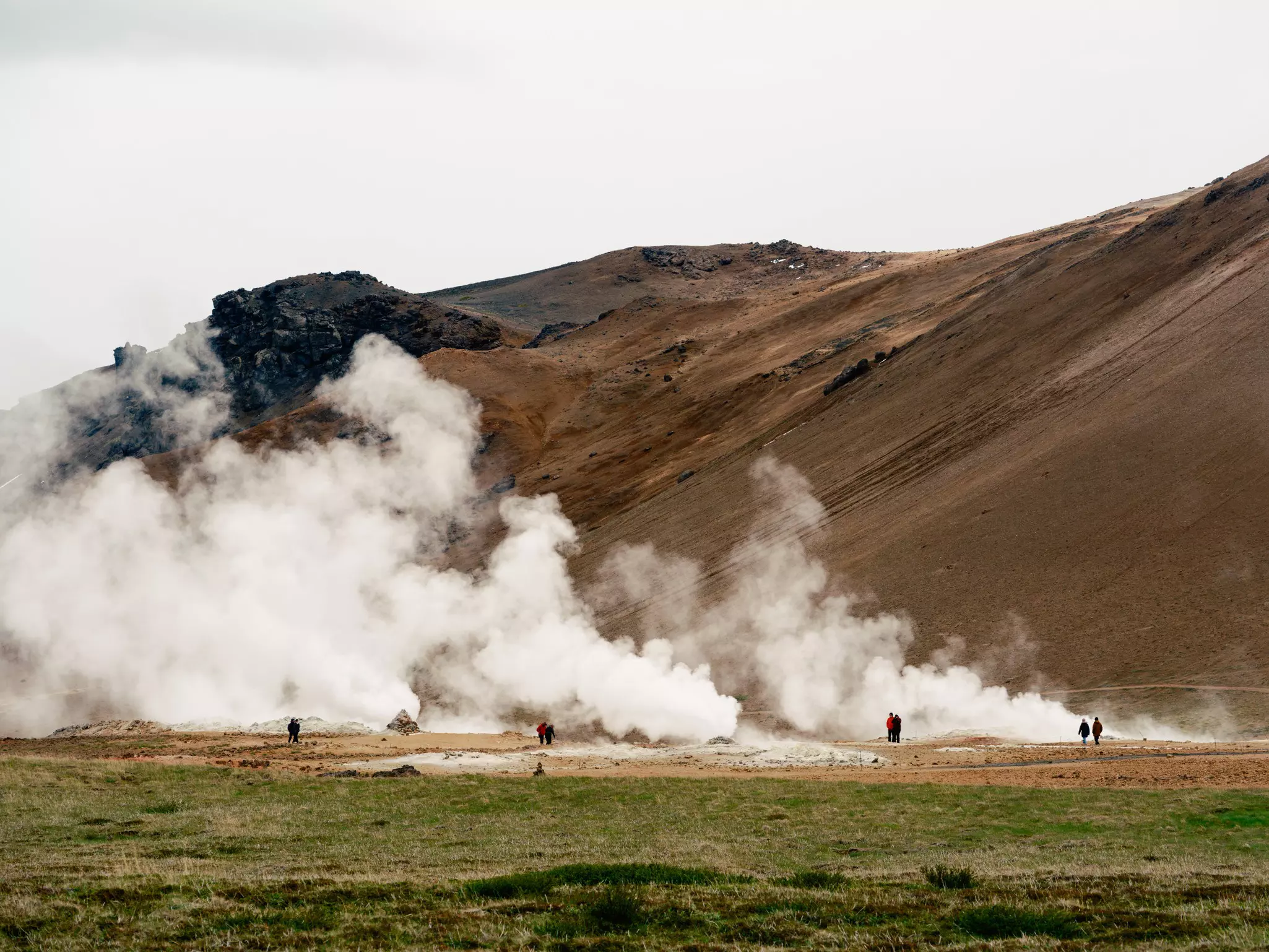 People walking near large clouds of steam rising from the ground with a dirt- and rock-covered mountain in the distance on an overcast day.
