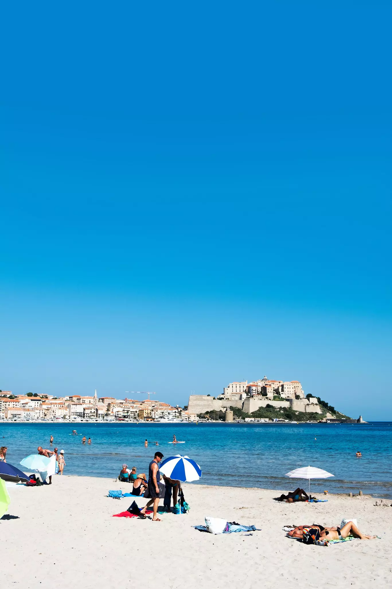 People relaxing on a public white-sand beach near a fortified city