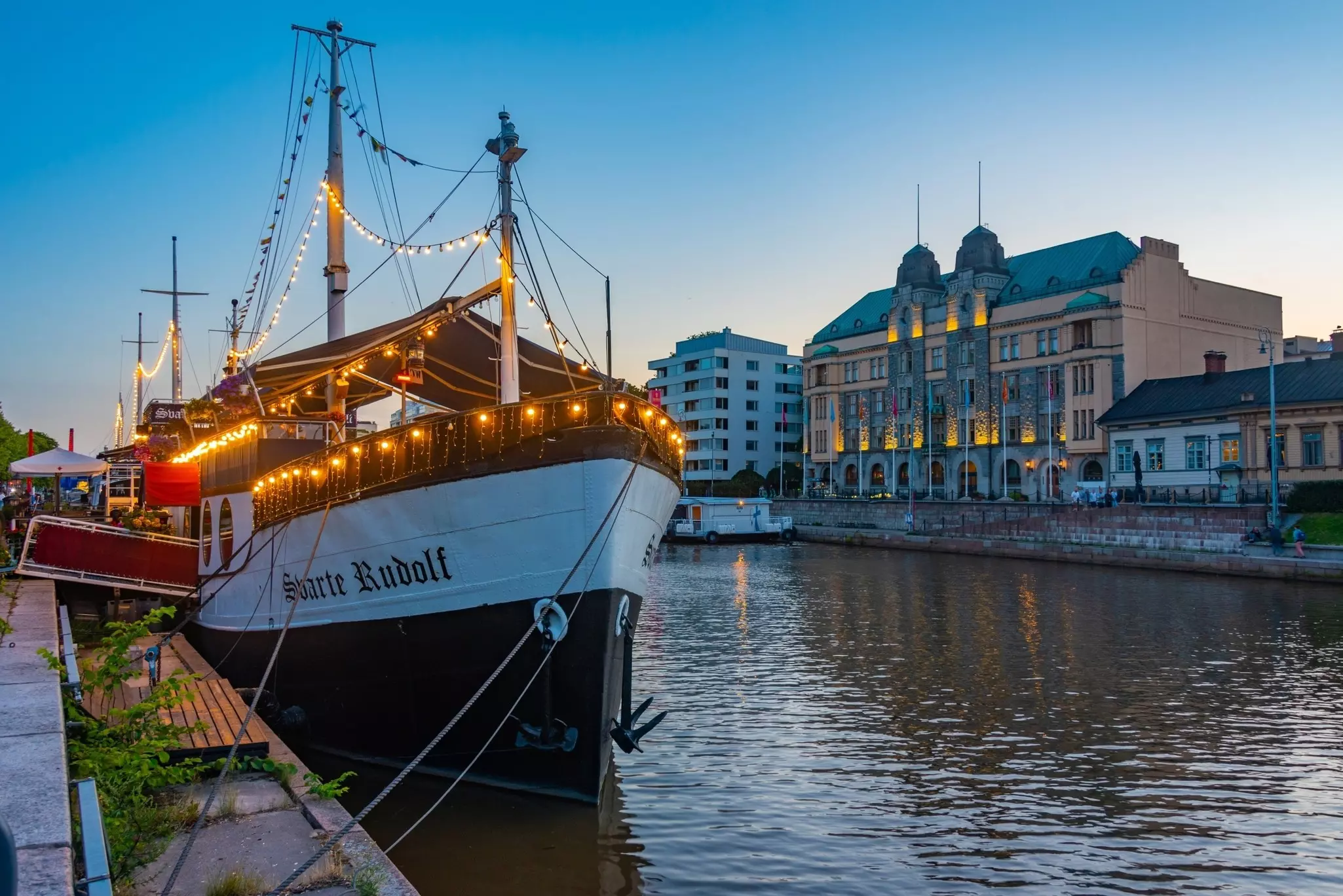 A boat adorned with strings of lights is moored by a city riverbank at dusk.