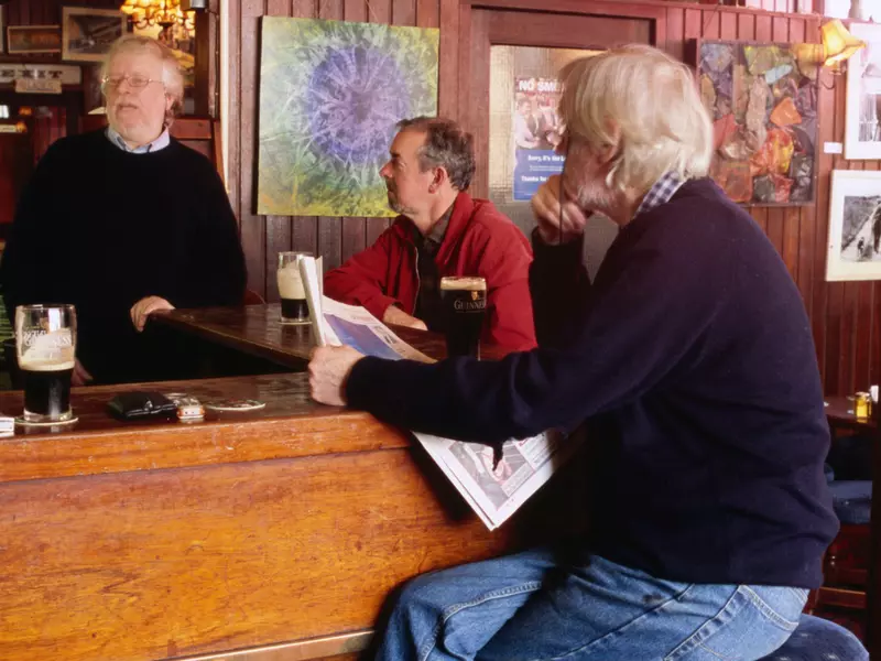 Three men sit at a bar with newspapers and pits of Guinness at the bar in front of them