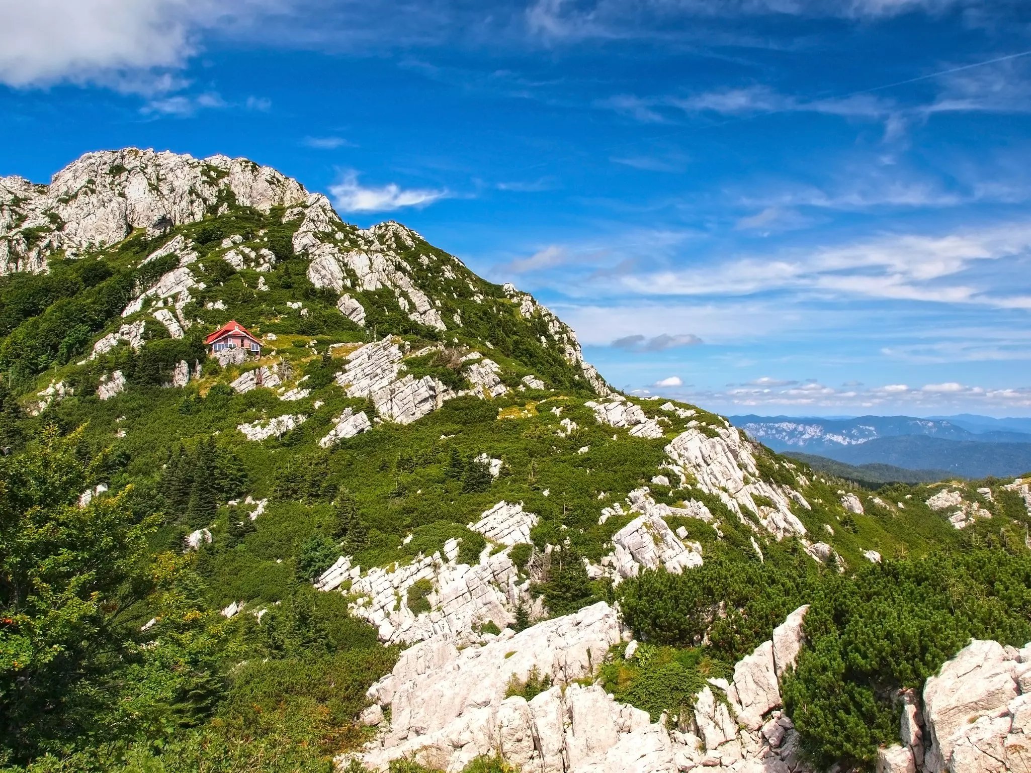 A single small cabin is set on a steep green mountain with white rocky outcrops under blue sky with white clouds.