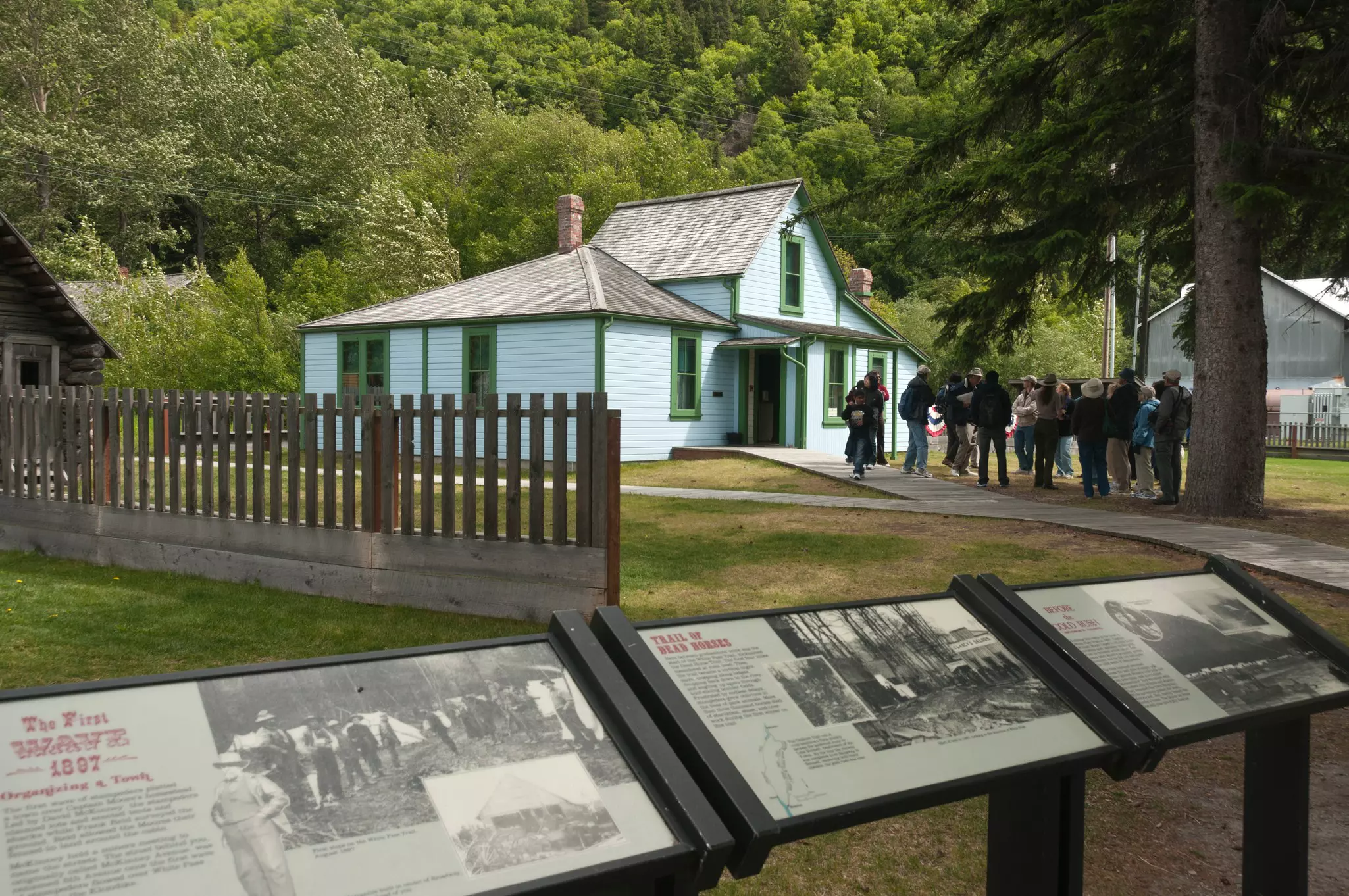 A ranger gives a talk to a group of tourists outside a small wooden house. Interpretive boards stand nearby sharing further information. 